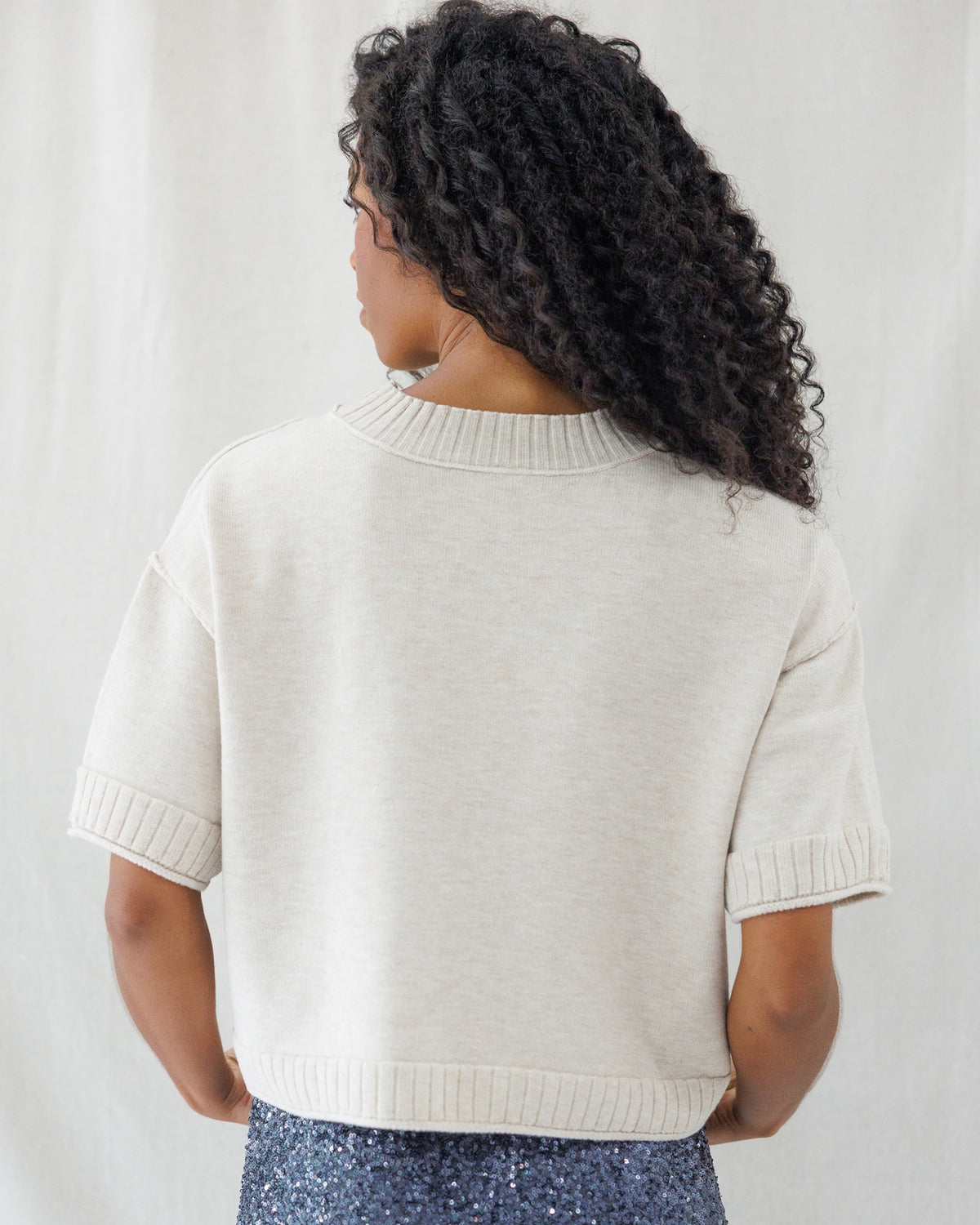 Back view highlighting the clean, loose fit of the beige knit top, detailed with exposed drop-shoulder seams, a thick ribbed collar, and matching ribbed hem