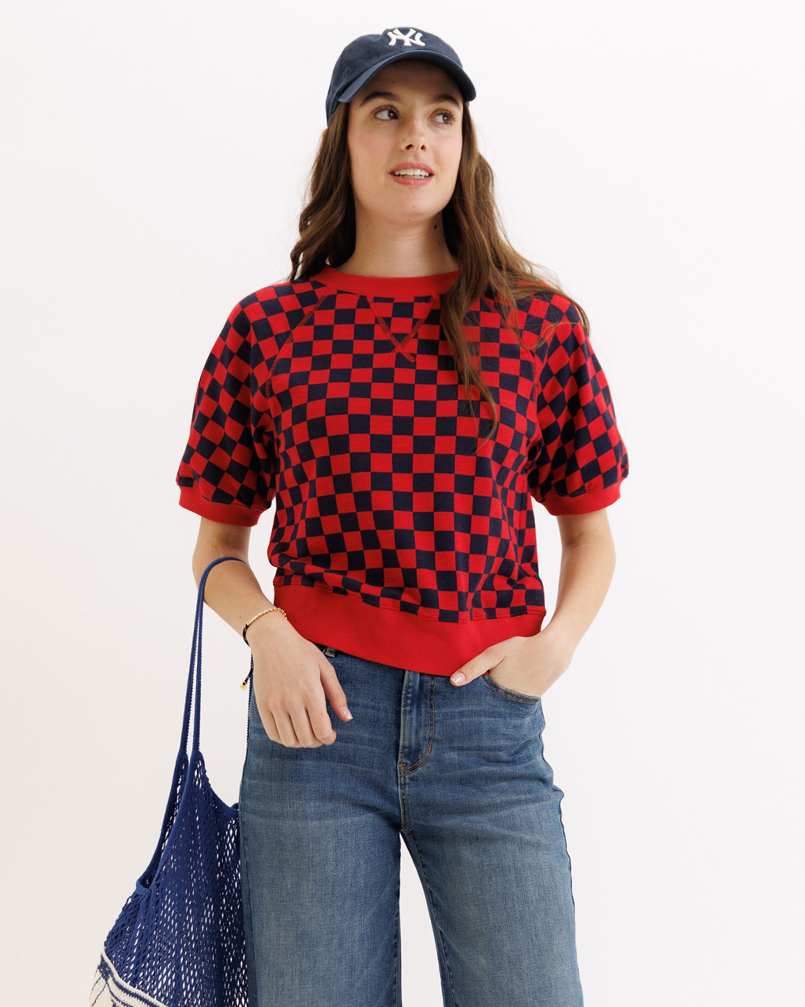 Person wearing a red and black checkered sweater with a blue cap and jeans, holding a blue bag on a white background.