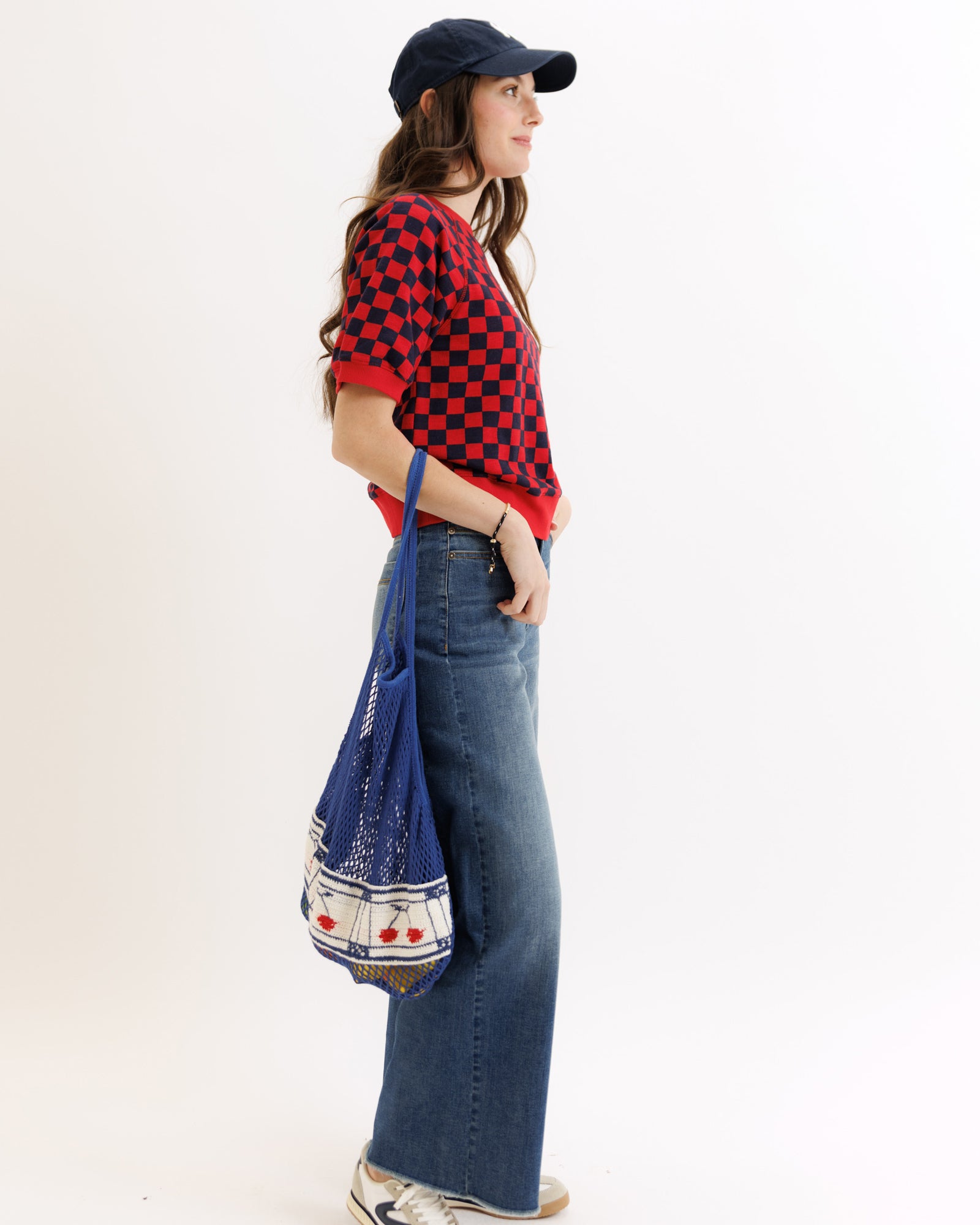 Person wearing a red checkered shirt, blue jeans, and a navy cap, holding a blue bag with cherry design.