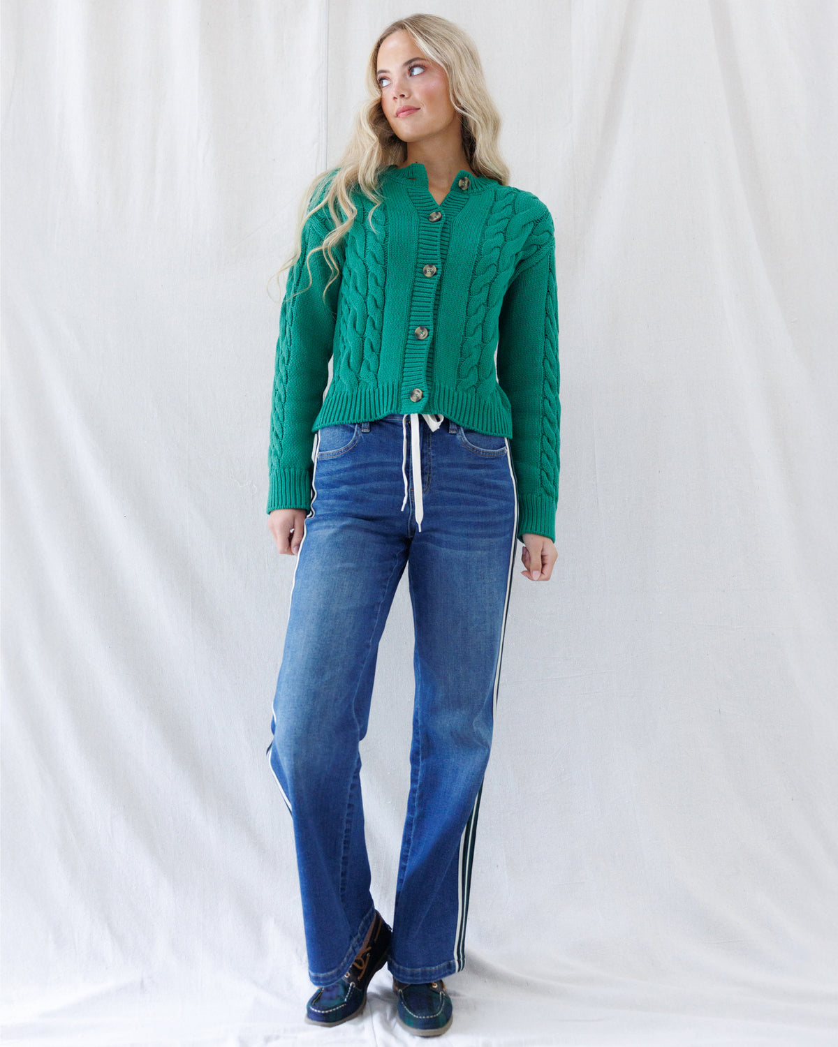 A woman modeling a bright green cable-knit cropped cardigan sweater with blue jeans featuring white side stripes, posed against a white fabric backdrop.