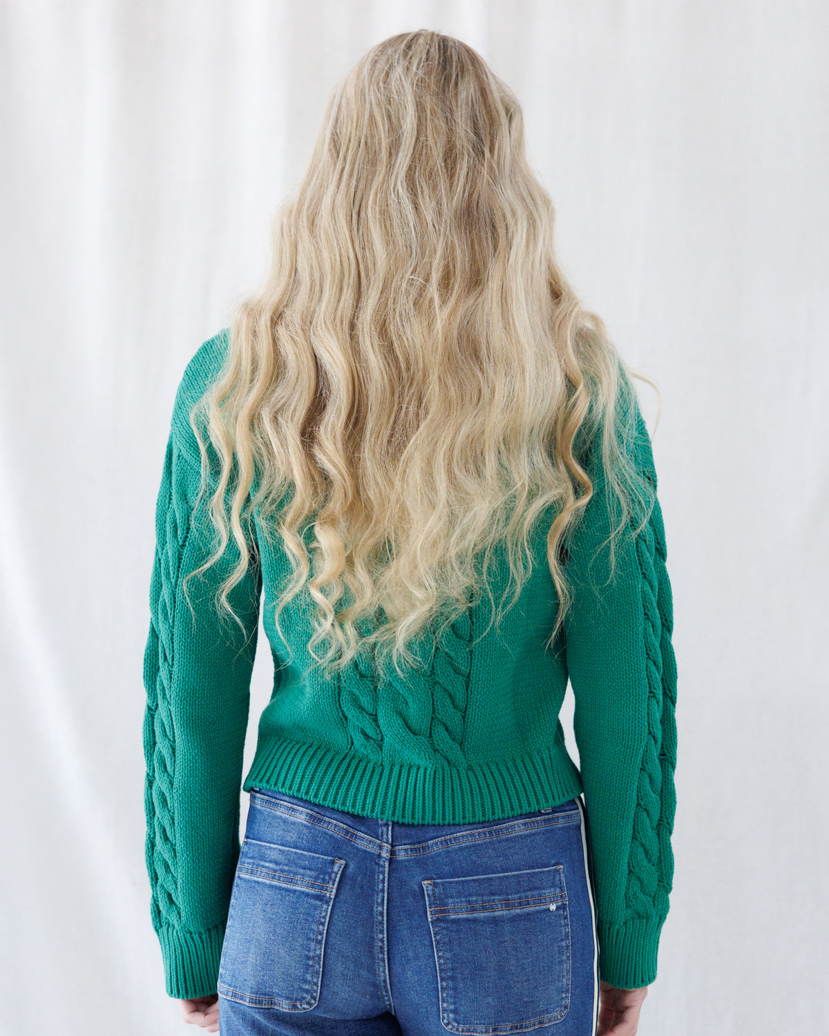 A woman viewed from behind wearing a green cable-knit sweater and blue jeans, standing against a white curtain background.