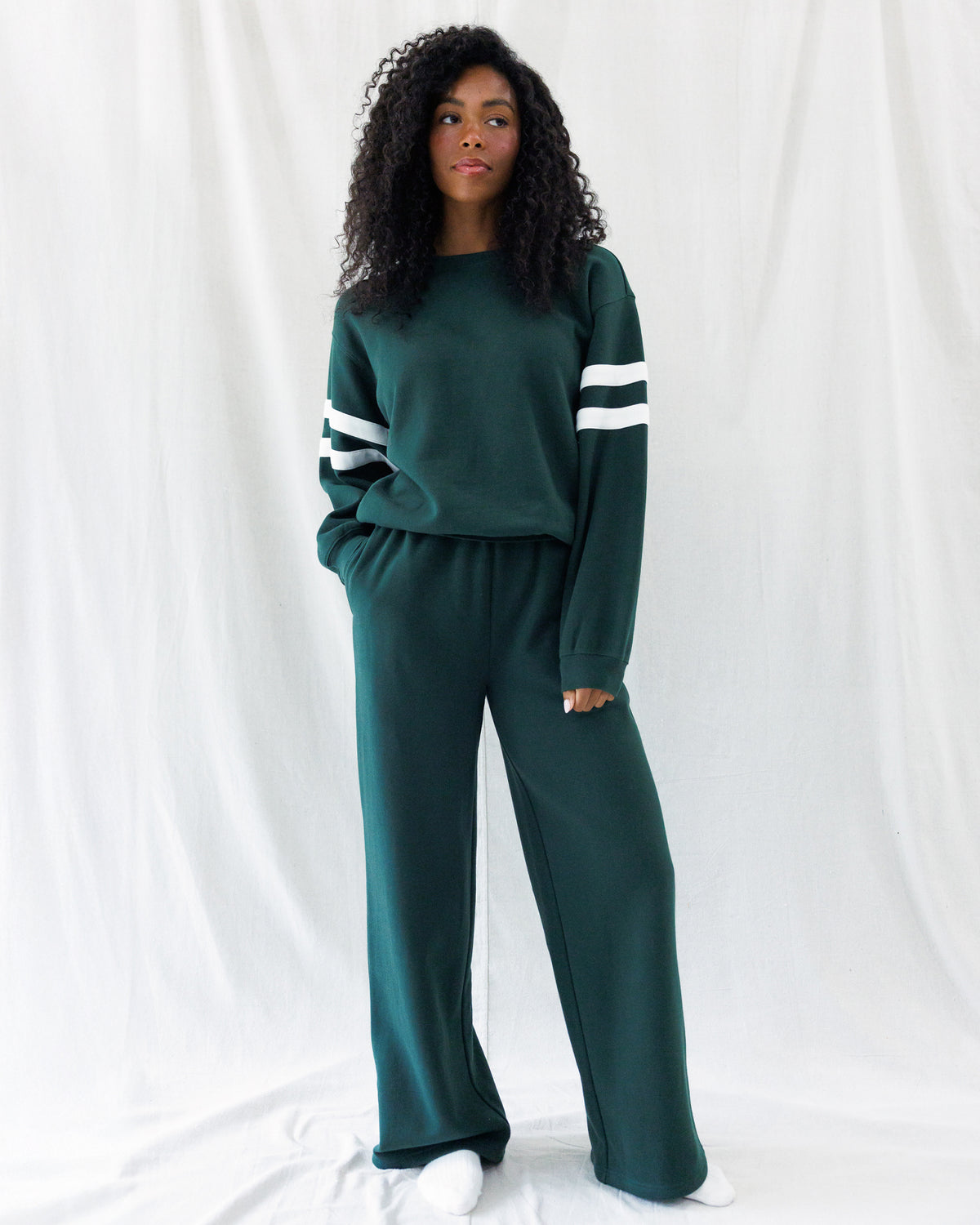 A woman modeling a dark green two-piece lounge set consisting of a long-sleeve sweatshirt with white stripes on the sleeves and matching wide-leg sweatpants, set against a white draped fabric background.