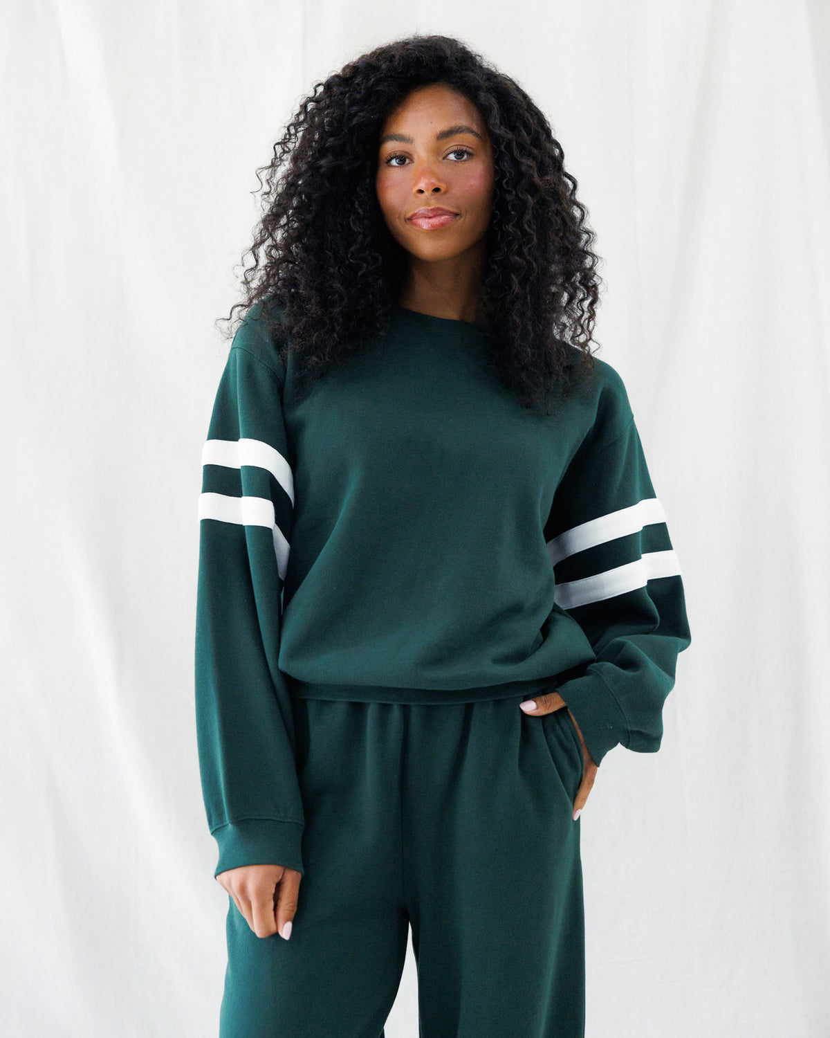 A woman with long, curly black hair wearing a dark green sweatshirt and matching sweatpants with white stripes on the sleeves, standing against a white fabric backdrop.