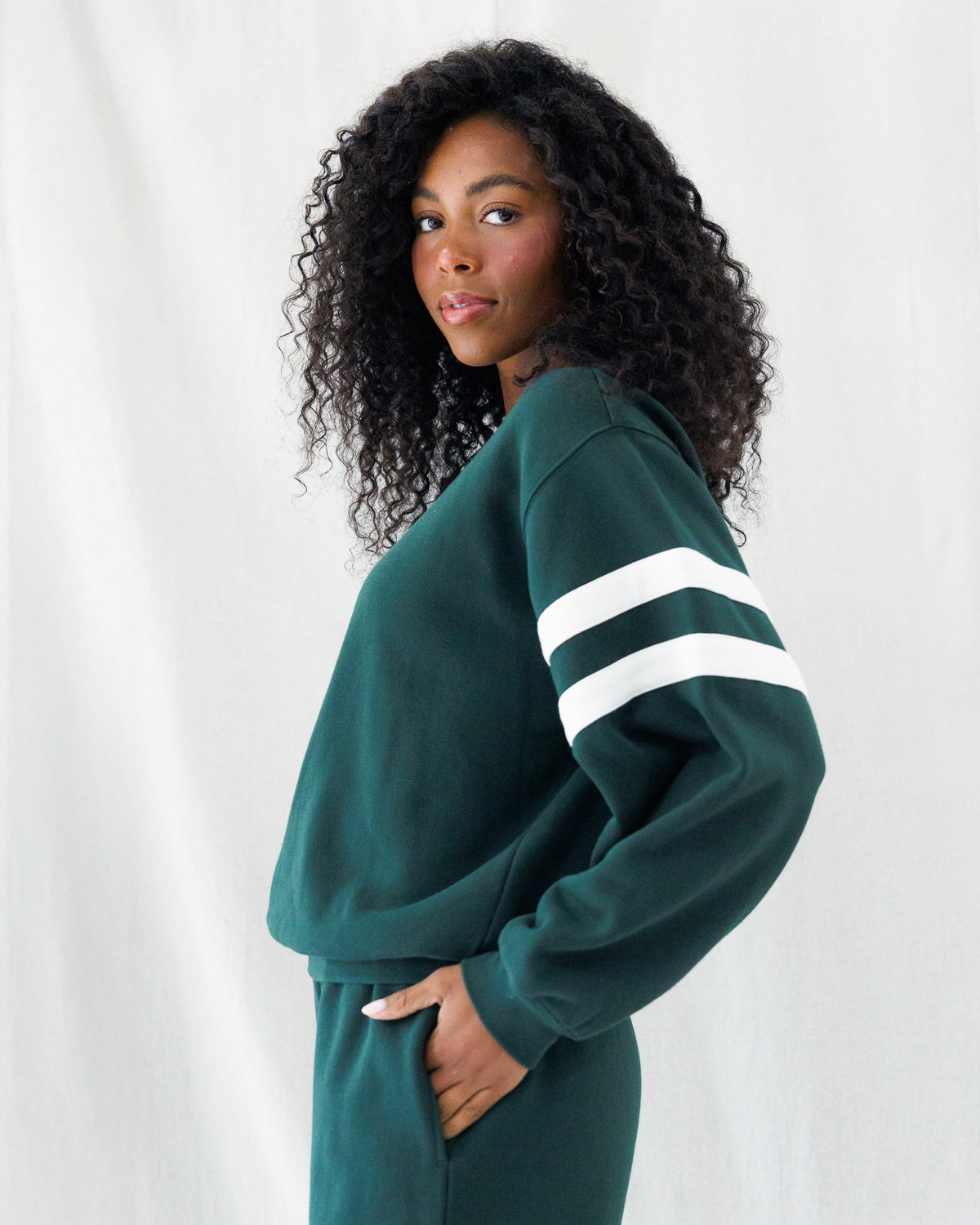 A woman with curly black hair wearing a dark green sweatshirt and matching sweatpants, featuring white athletic stripes on the sleeves, posed against a white draped fabric background.