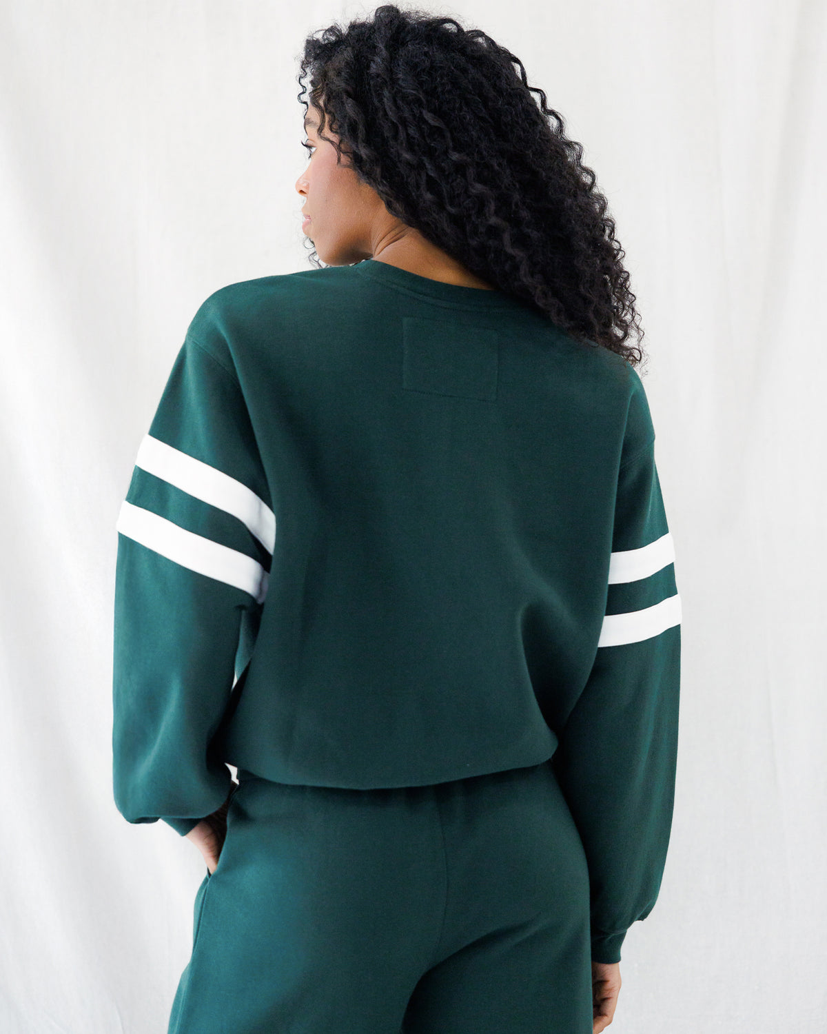 A woman shown from the back wearing a dark green cropped sweatshirt with white stripes on the sleeves and matching dark green sweatpants, against a white draped background.