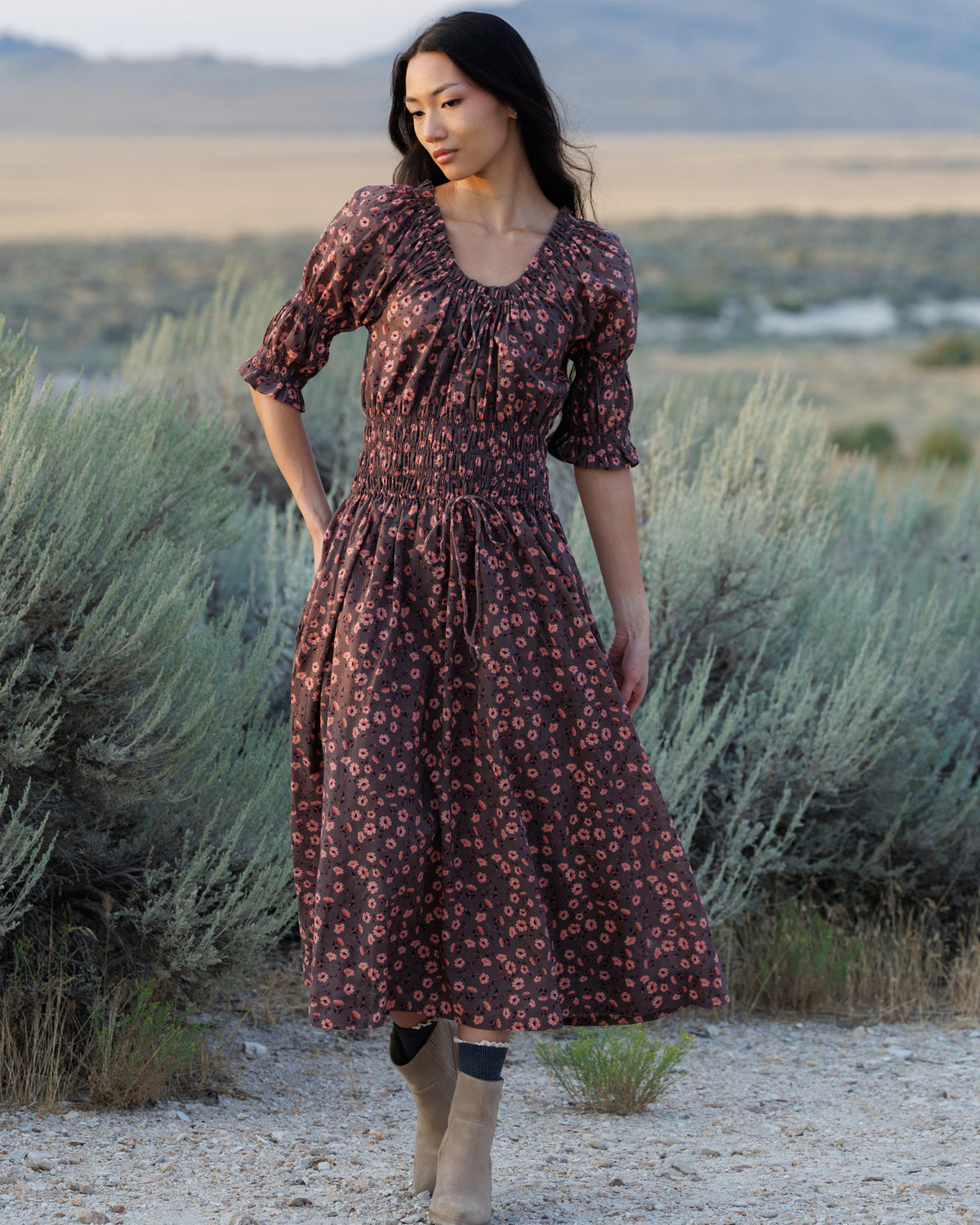 A woman wearing a dark brown midi dress with a small pink floral pattern stands in a desert landscape with sagebrush and mountains in the background.