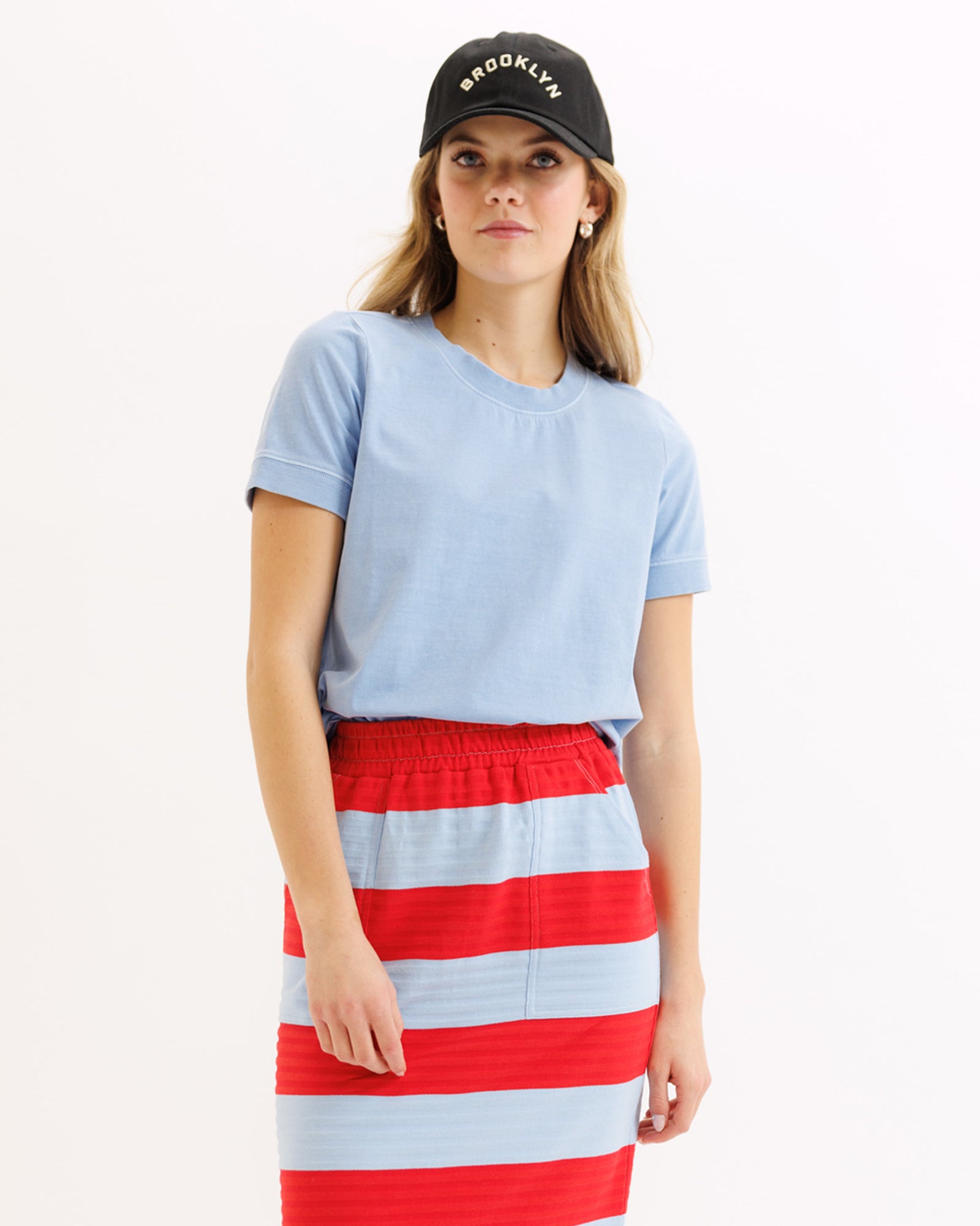 Woman wearing a light blue t-shirt and red and white striped skirt with a black cap.