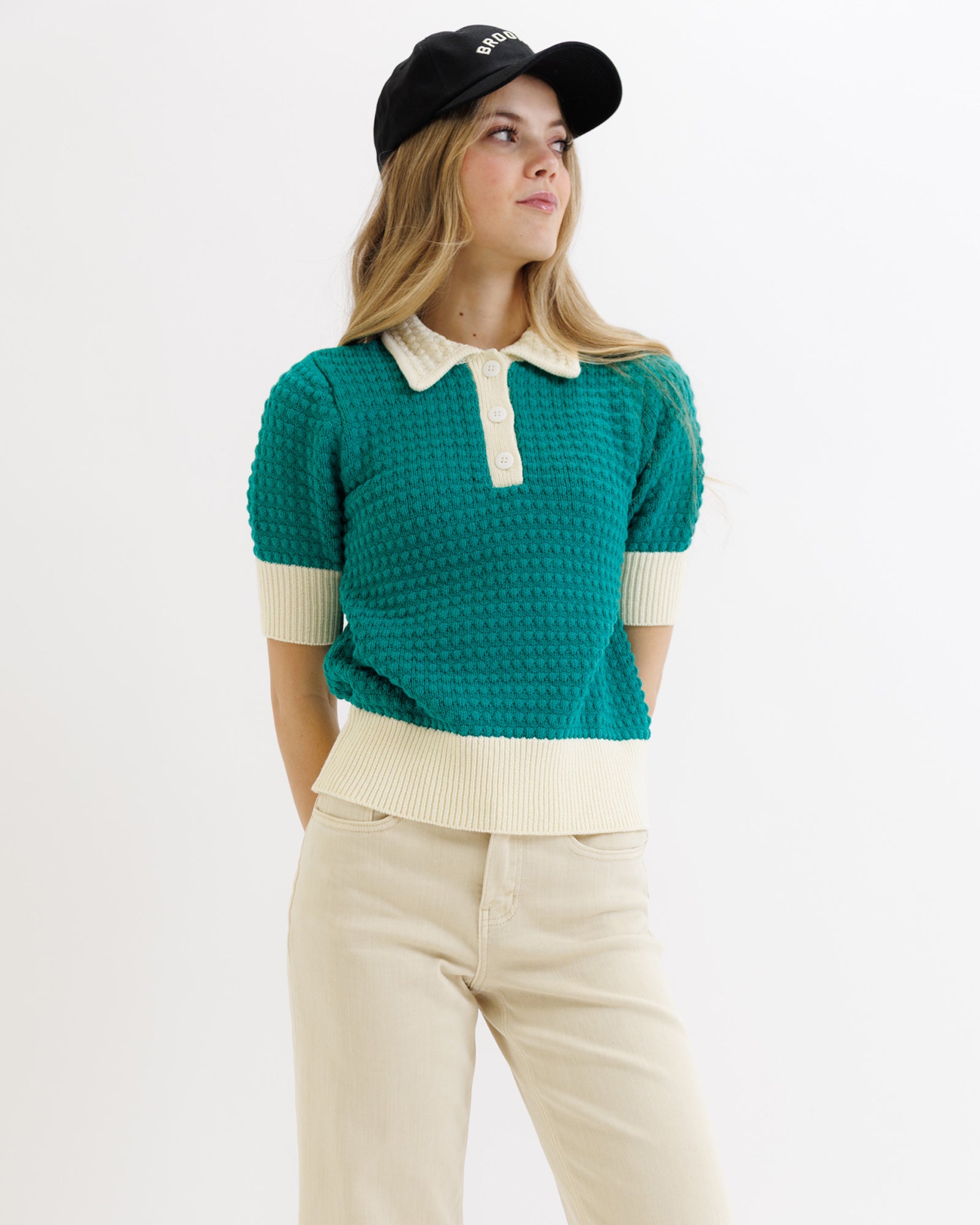 Person wearing a green textured sweater with white collar and cuffs, beige pants, and black cap on a light gray background