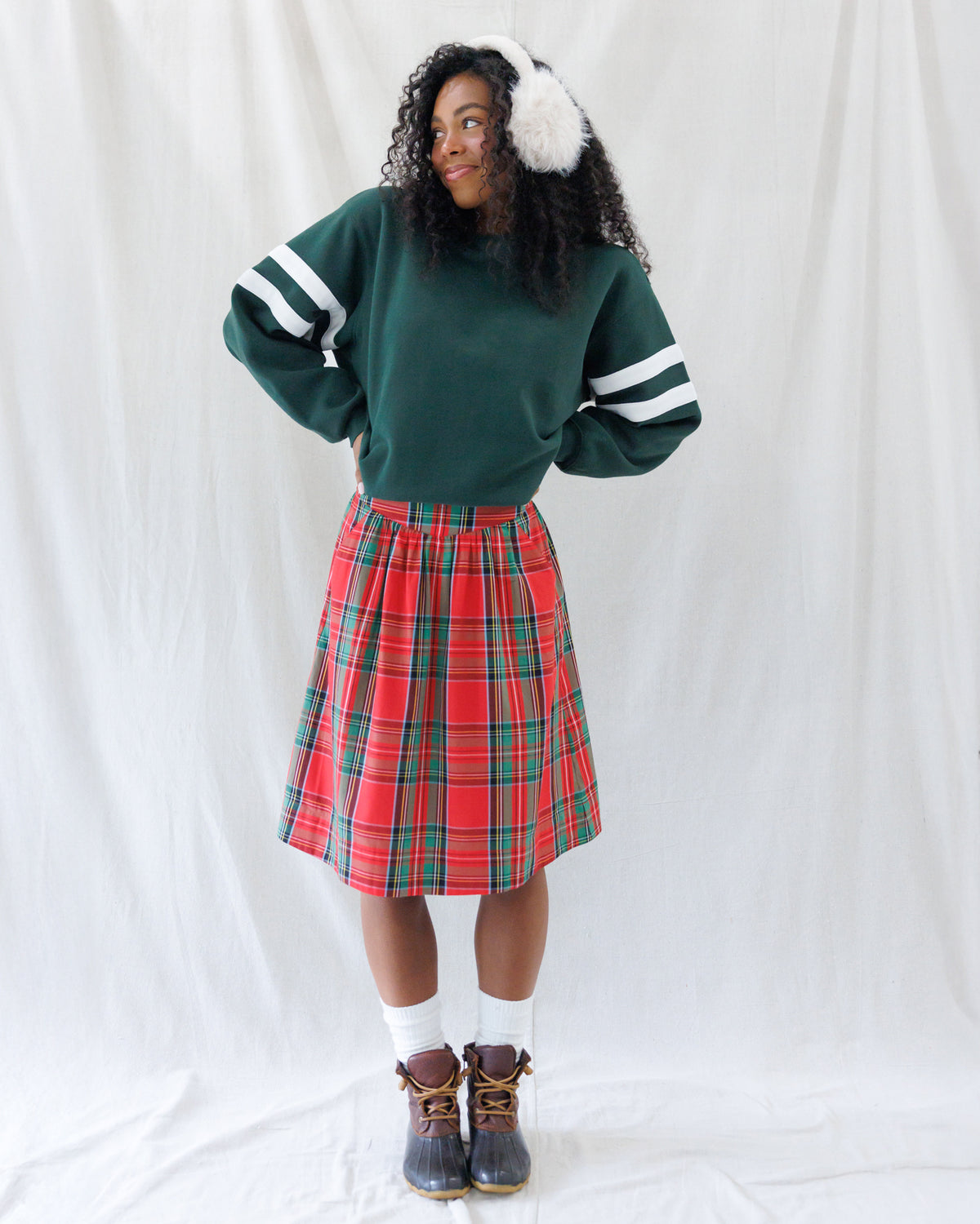 A woman stands against a white fabric backdrop wearing a dark green crewneck sweatshirt with white stripes on the sleeves, a red and green plaid pleated skirt, white socks, and brown boots. She is also wearing white faux-fur earmuffs.