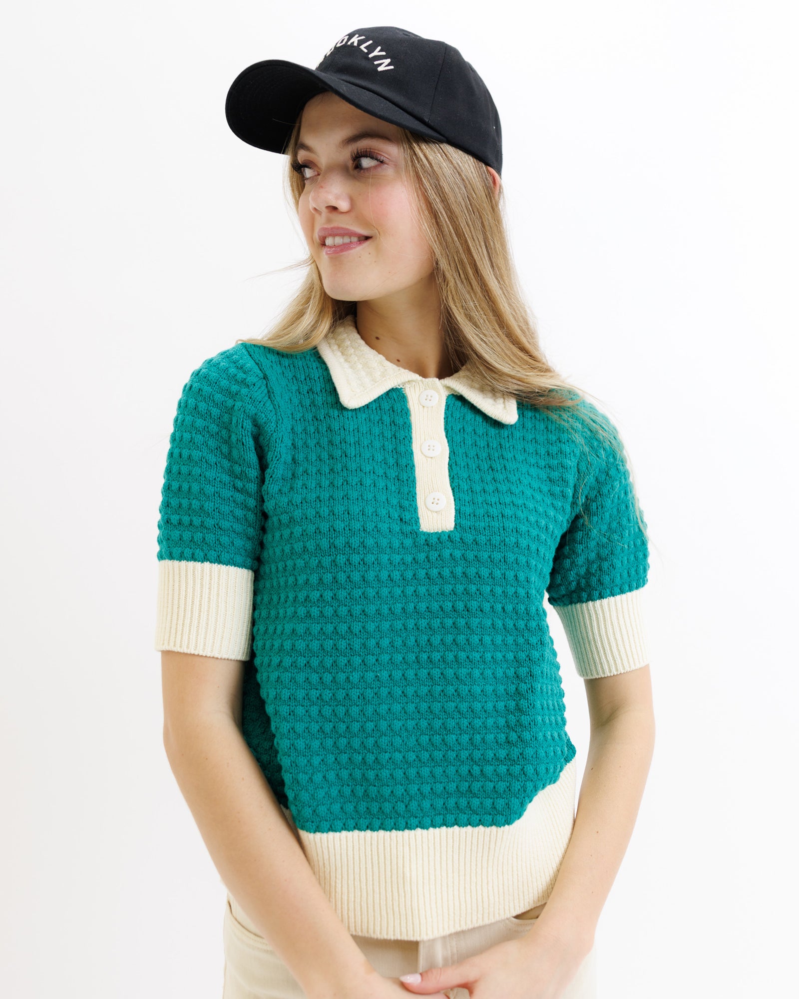 Woman wearing a teal knitted polo shirt with white collar and cuffs, and a black cap.
