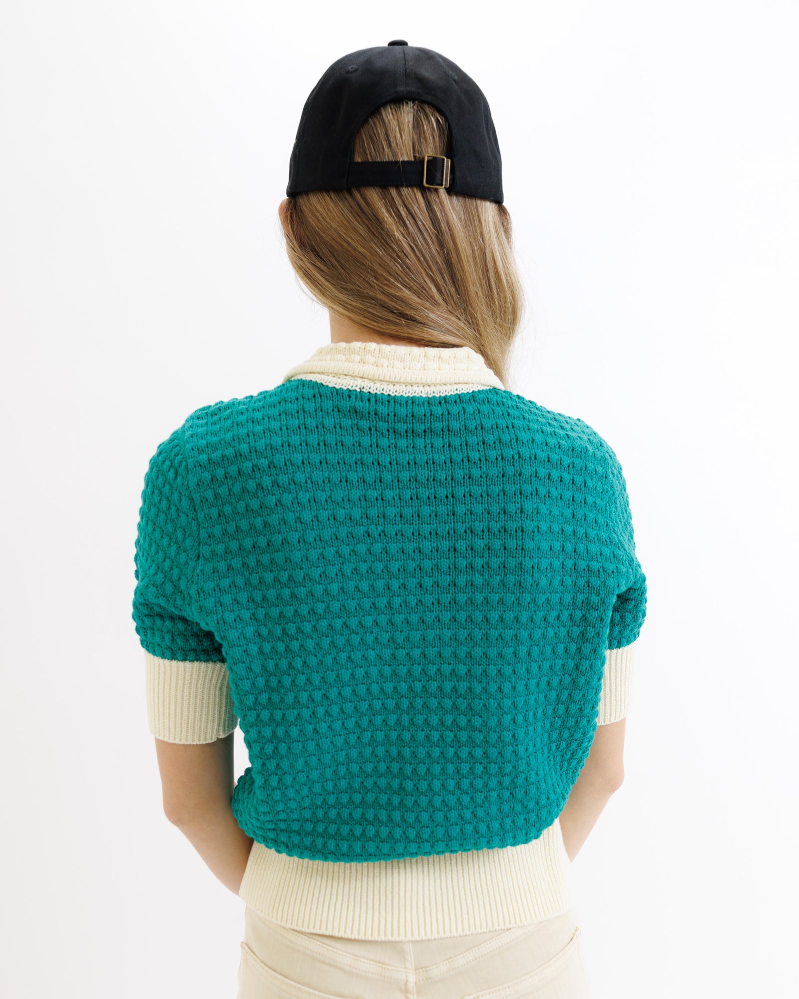 Person wearing a teal knitted cardigan and black cap on a white background