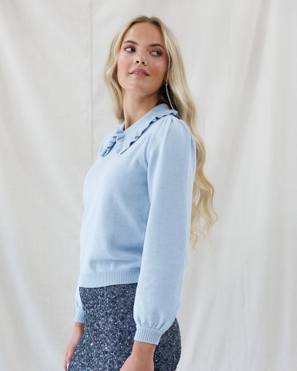 A blonde woman wearing a light blue long-sleeve knit sweater with a ruffled collar and dark patterned pants, standing against a draped white fabric backdrop.