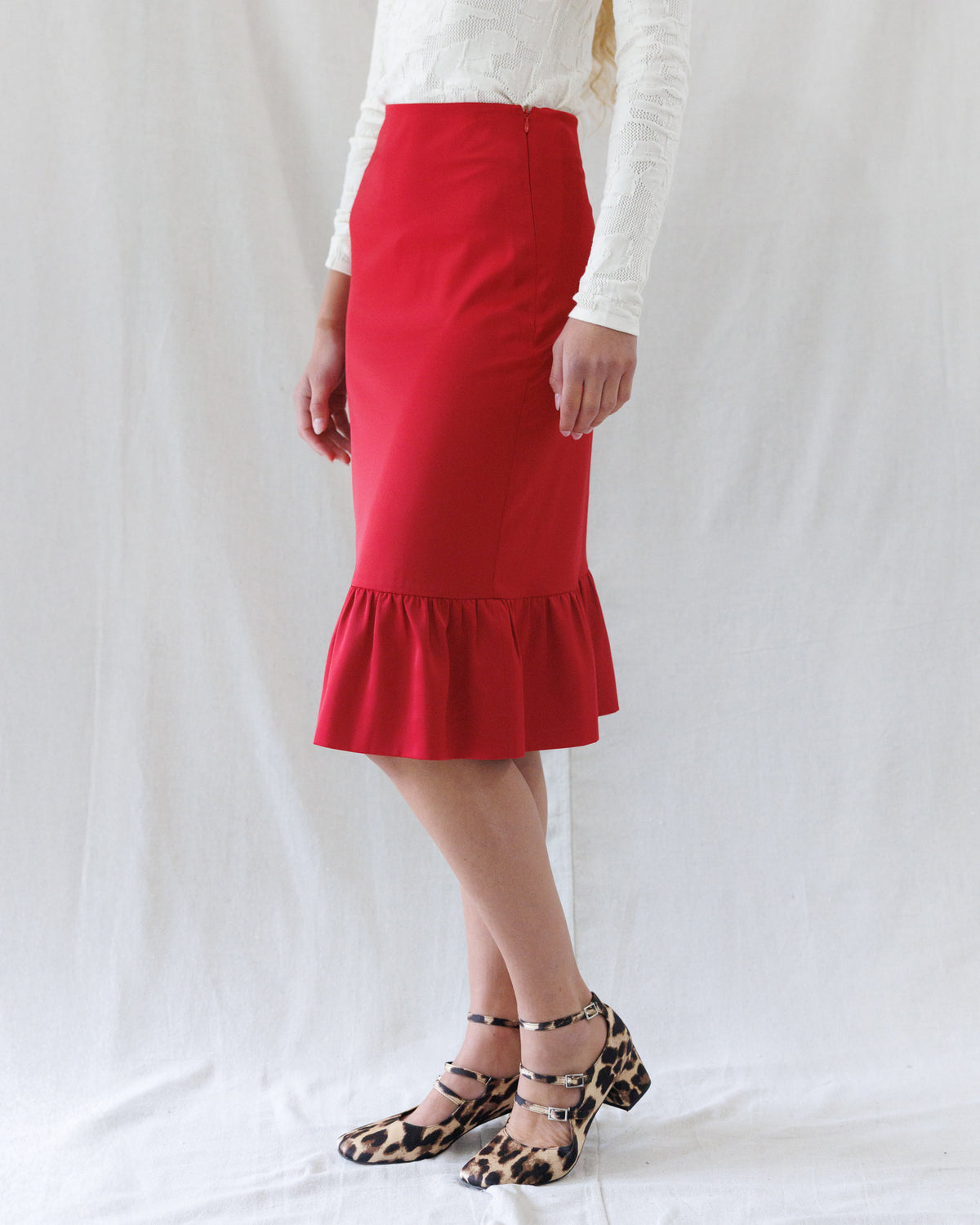 A model wearing a bright red knee-length pencil skirt with a ruffled hem, paired with a white long-sleeve top and leopard print heels, standing against a white fabric backdrop.