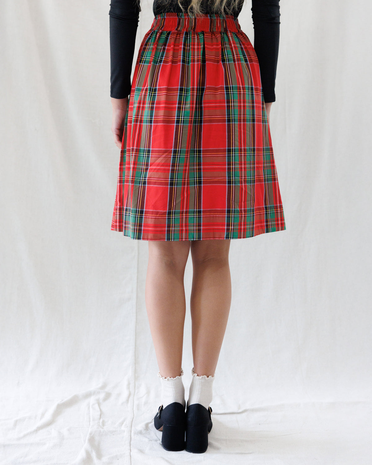A person shown from the back wearing a red tartan plaid skirt with green and black stripes, paired with a black long-sleeve top, white socks, and black shoes, standing against a white fabric backdrop.