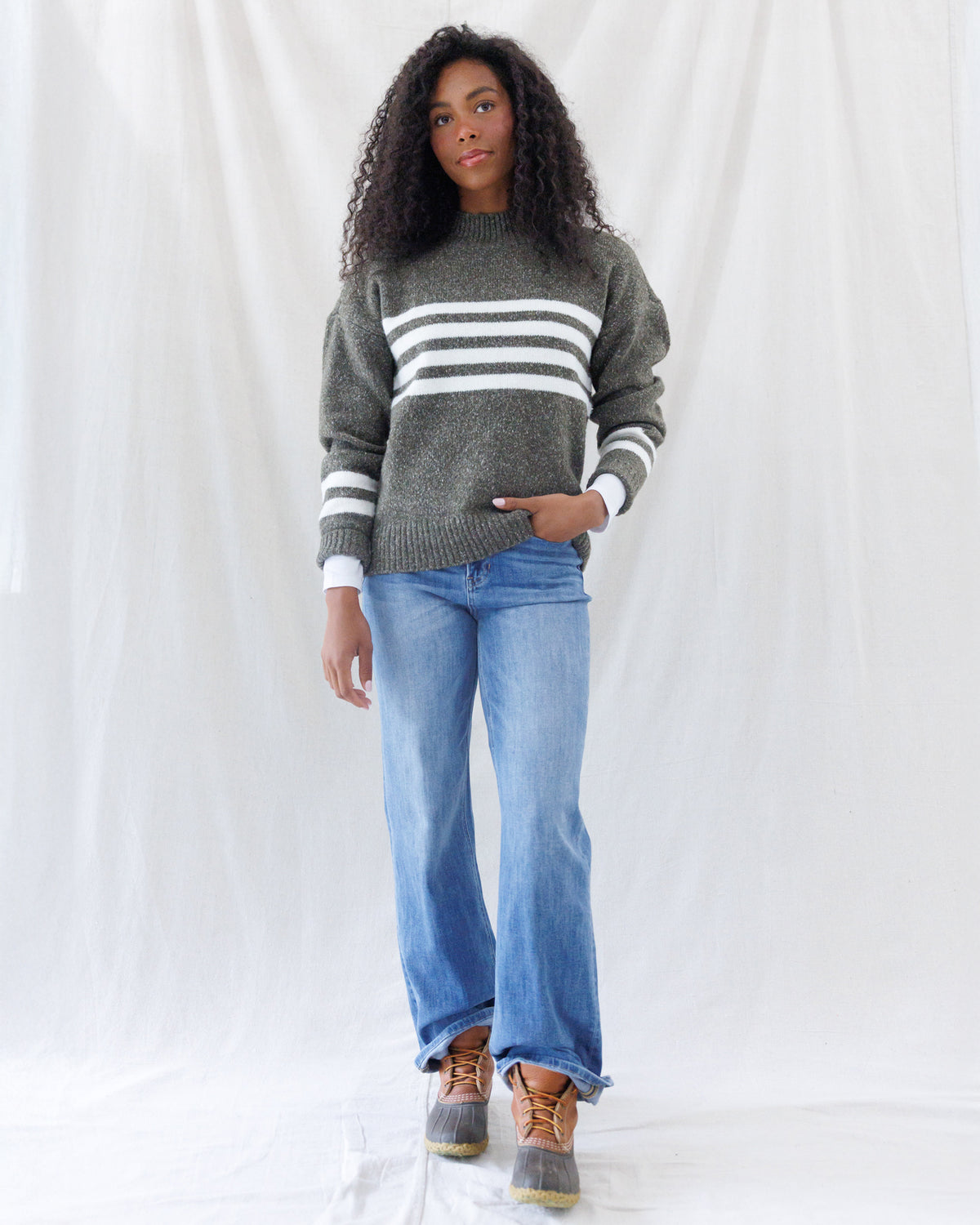 Model wearing a relaxed-fit olive marled knit sweater featuring bold white chest stripes, drop shoulders, and ribbed trim, styled untucked over light-wash flare jeans.
