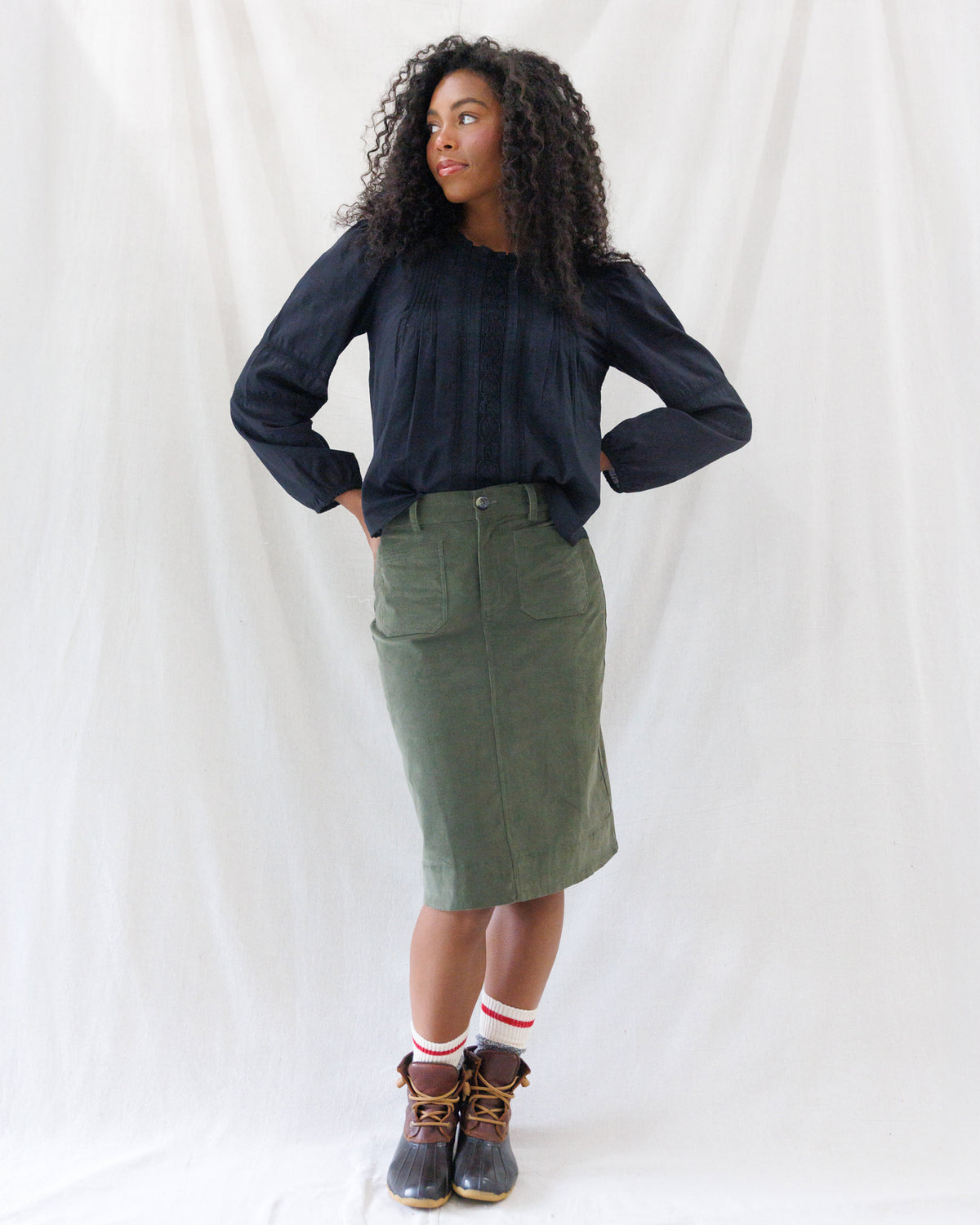 Model wearing the olive corduroy knee-length skirt styled with a dark button-up peasant blouse loosely tucked at the waist.