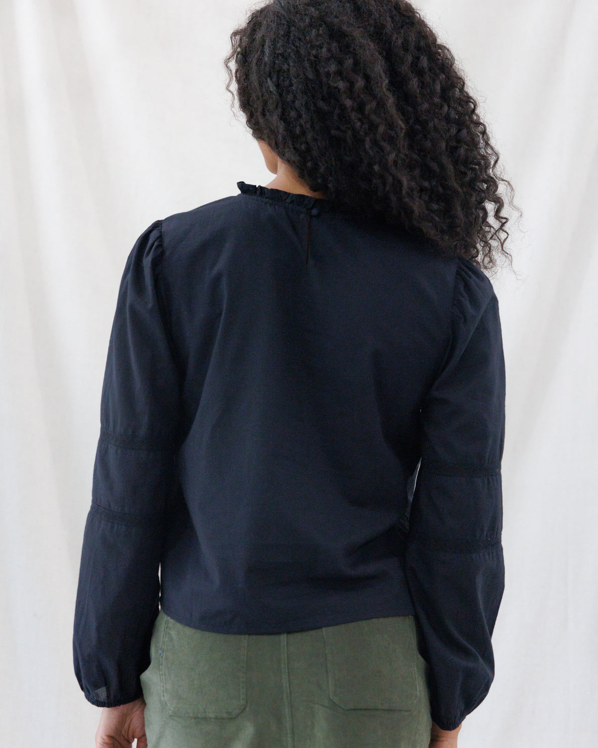 Back view highlighting the clean, easy-fitting silhouette of the black peasant top with a delicate ruffle-trimmed collar, keyhole loop closure, and hip-length hem.