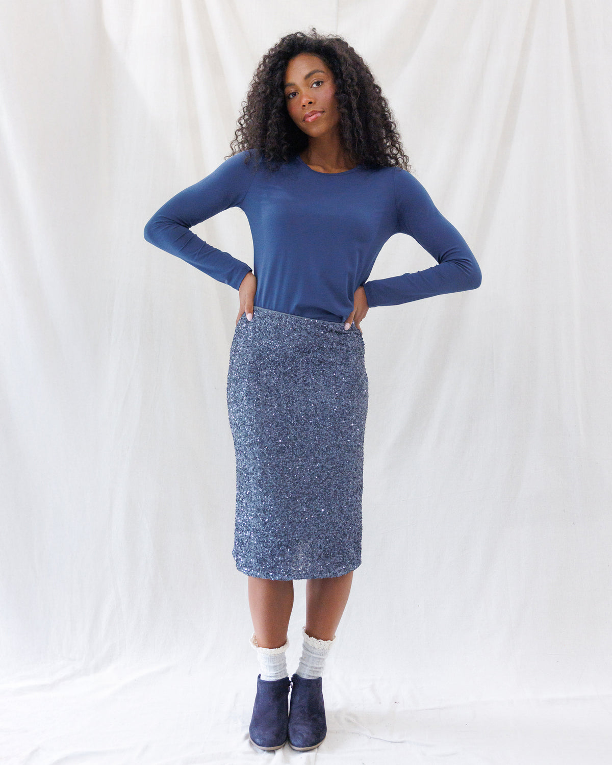 A woman modeling a blue long-sleeve crewneck top and a sparkly blue sequined pencil skirt, paired with white socks and navy blue boots, standing against a white fabric backdrop.
