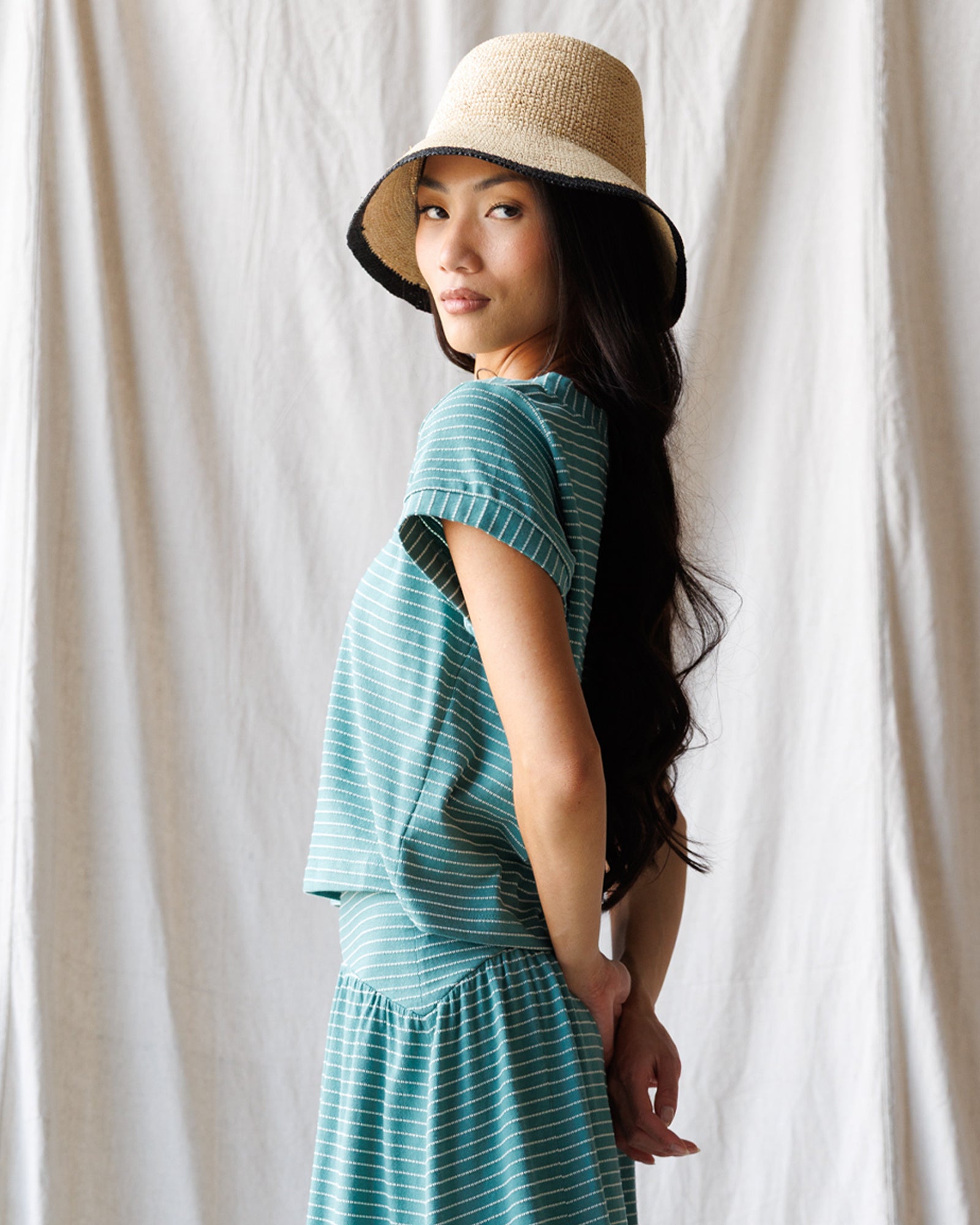 Woman wearing a teal dress and straw hat against a white curtain background