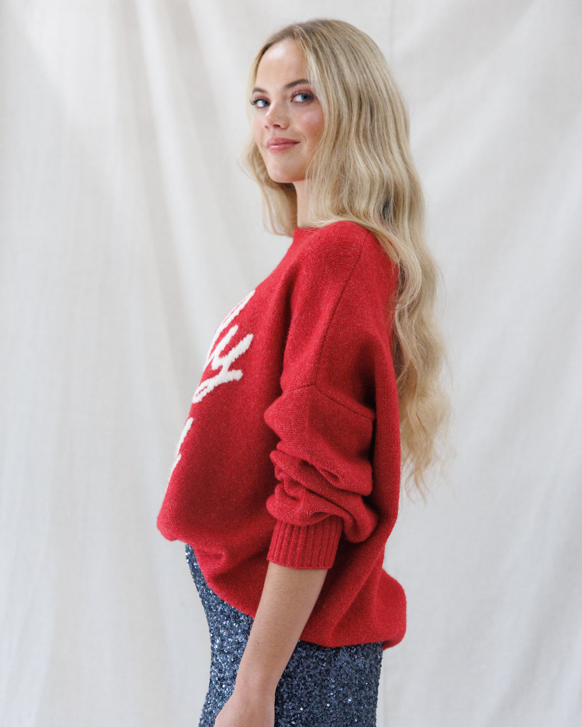 A blonde woman wearing a red knit sweater with a white graphic design on the front, paired with sparkly blue pants, posing against a white draped fabric background.