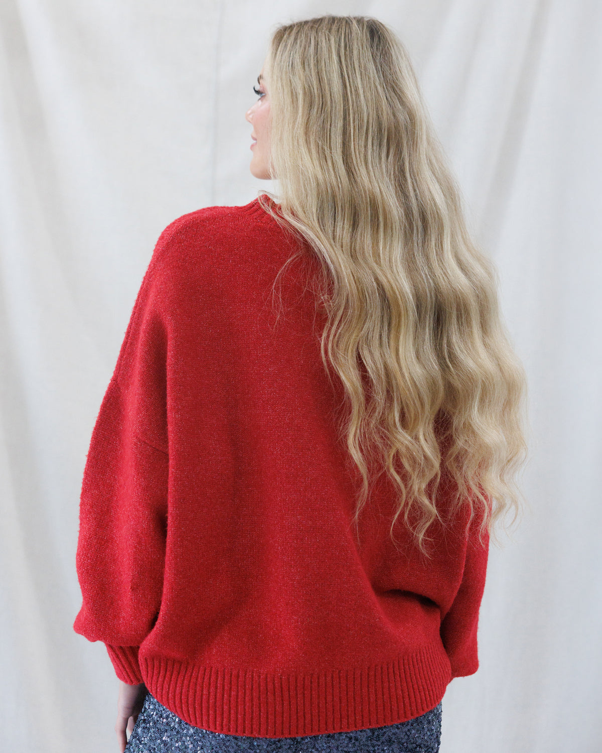 A woman with long, wavy blonde hair is shown from behind wearing a bright red knit sweater with a crew neck and ribbed cuffs and hem, set against a draped white fabric background.