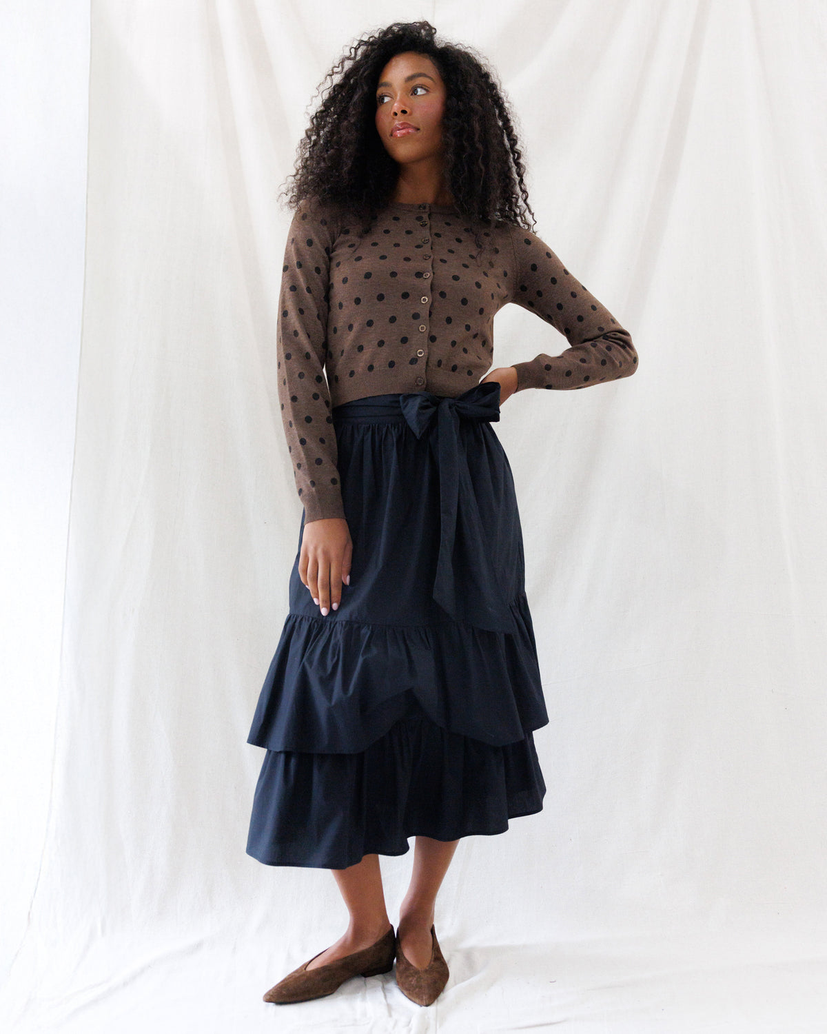 A-line black midi skirt featuring an optional matching self-tie sash at the waist and two full, gathered flounce tiers, styled with a brown polka-dot cardigan.