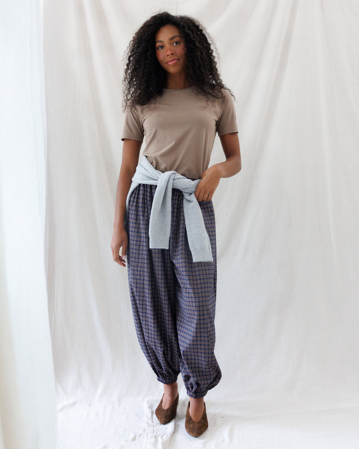 A woman stands against a white fabric backdrop wearing a taupe short-sleeve t-shirt, a light blue shirt tied at the waist, and dark blue and gray checkered harem pants with brown shoes.