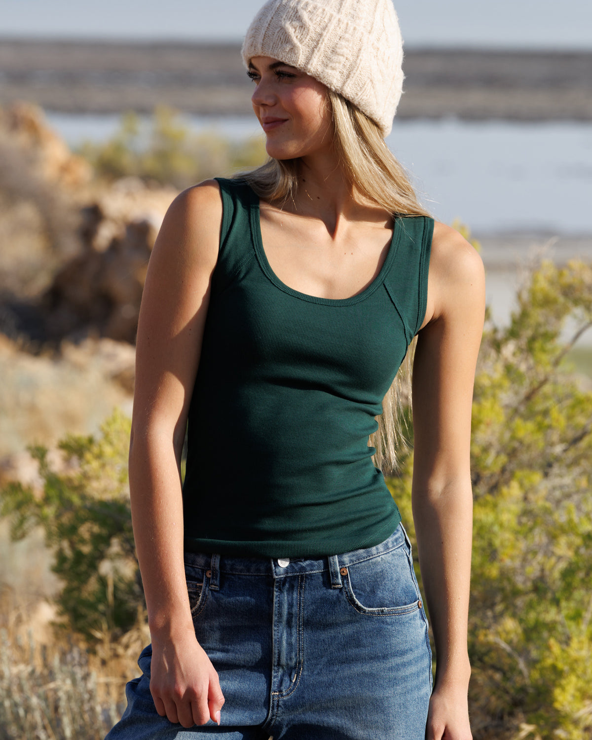 A woman wearing a dark green scoop-neck tank top, blue jeans, and a cream knit beanie, standing outdoors in a natural, rocky environment.