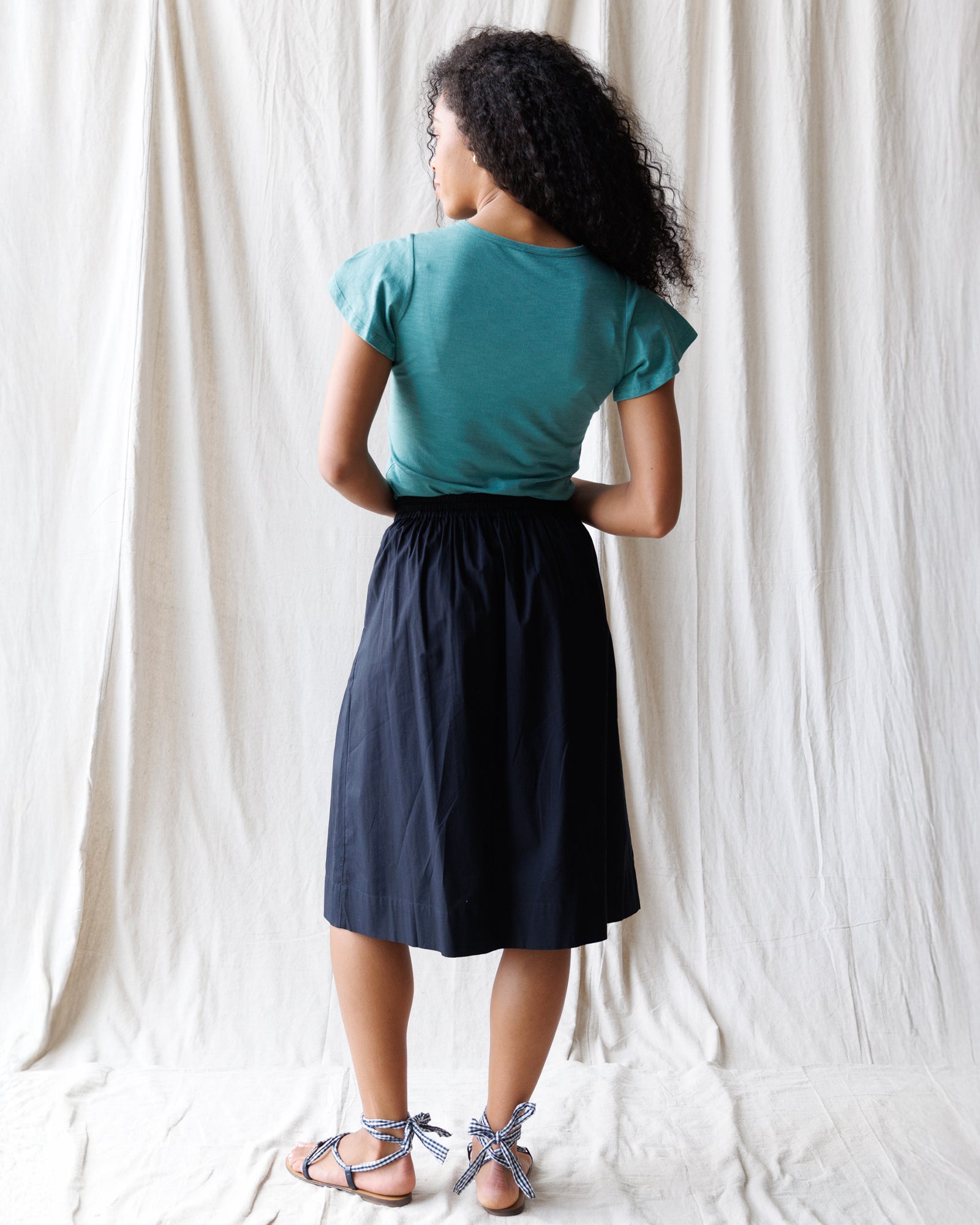 Person wearing a teal top and black skirt against a white curtain background