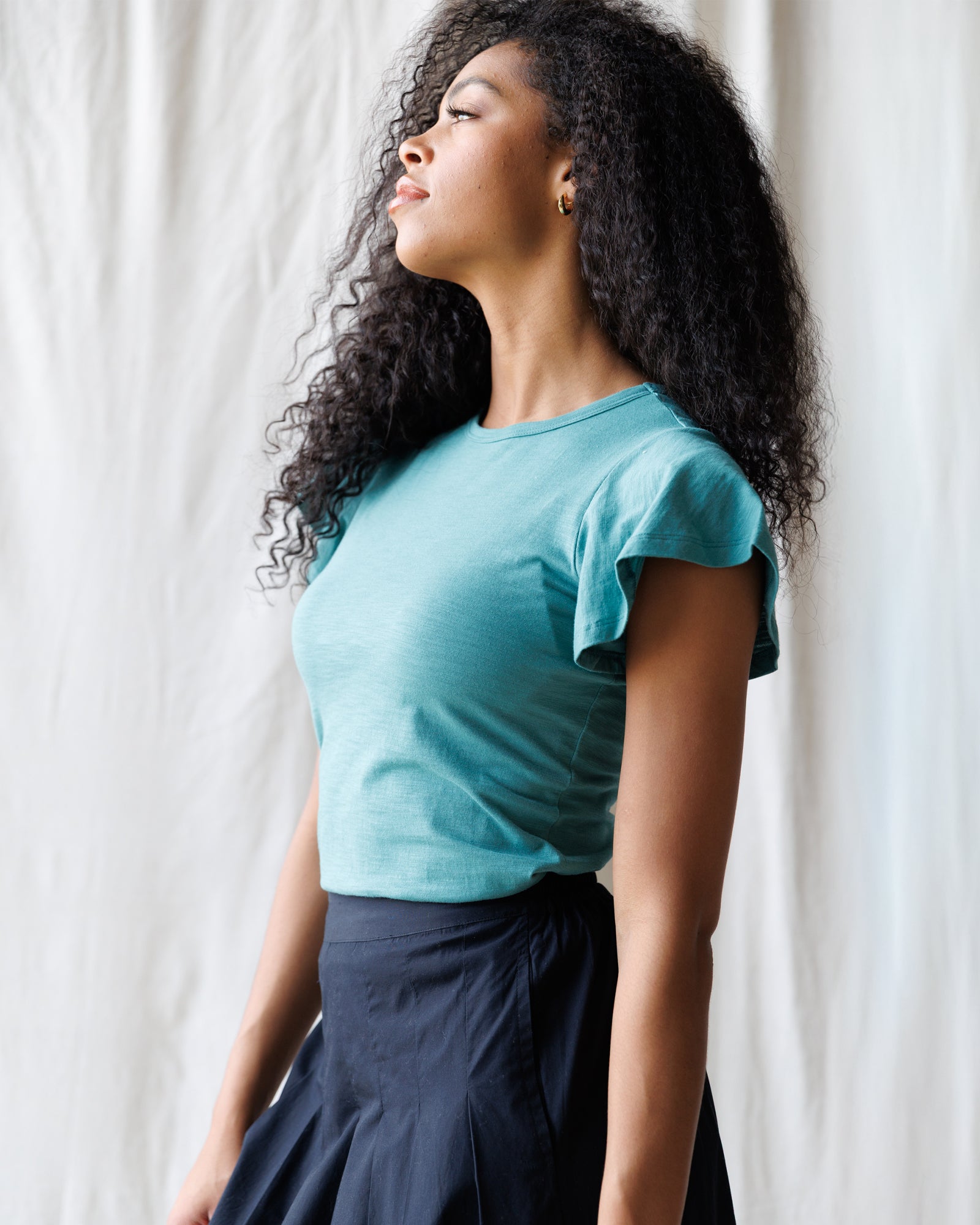 Woman wearing a teal top and black skirt against a white curtain background
