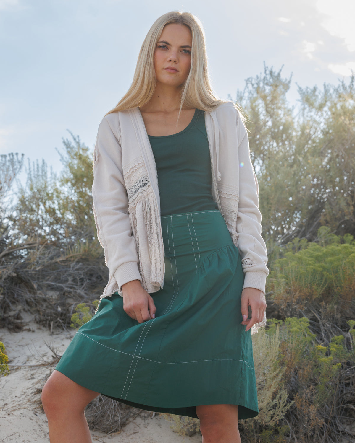A blonde woman wearing a cream cardigan, dark green tank top, and a dark green pleated skirt, standing outdoors in a sandy, brushy environment.