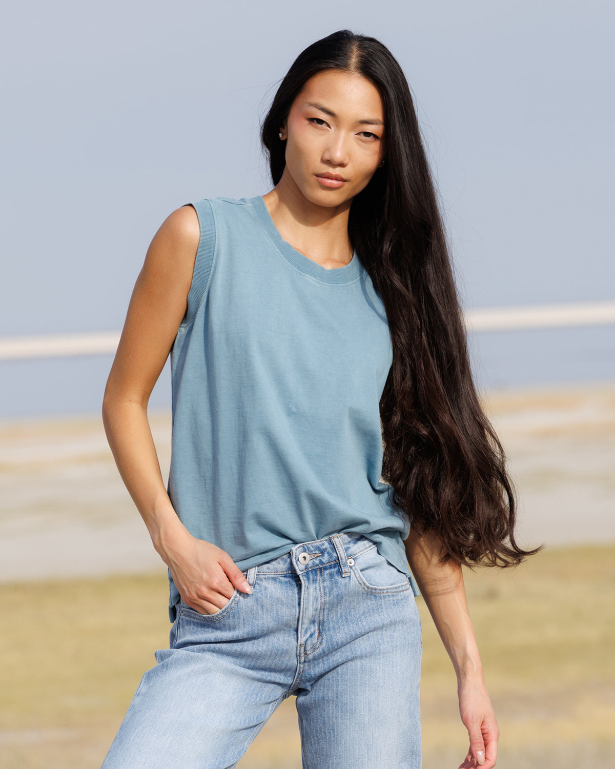 A woman with long dark hair wearing a light blue sleeveless crewneck top and light-wash blue jeans, standing outdoors against a soft-focus background of sky and grass.