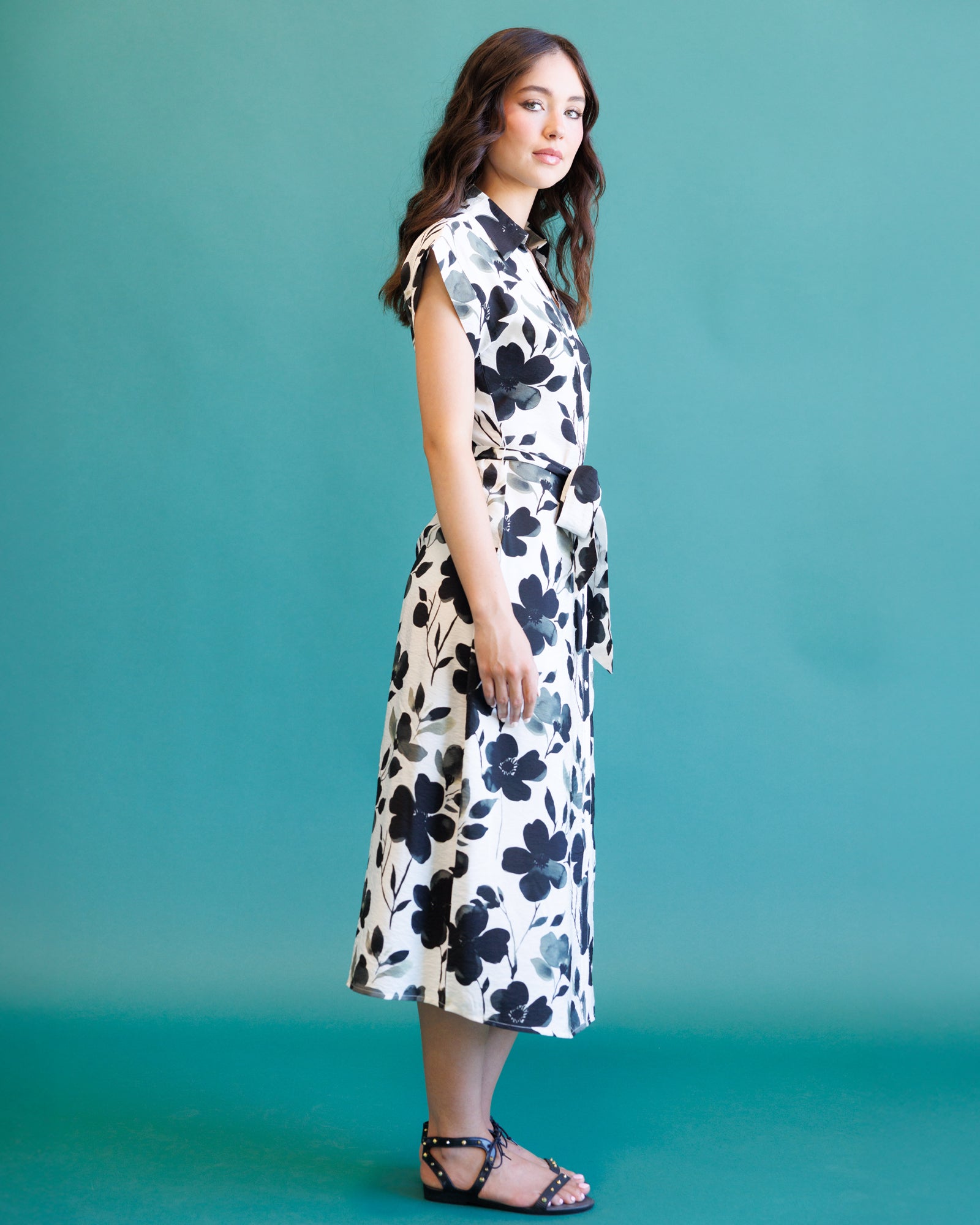 A woman modeling a sleeveless midi dress with a black floral print on a white background, standing against a solid teal studio backdrop.