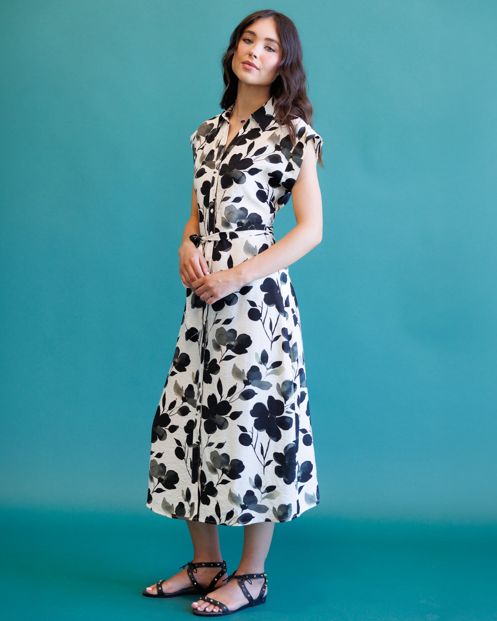A woman modeling a sleeveless midi dress with a black and gray floral print on a cream background, paired with black strappy sandals, set against a solid teal studio backdrop.