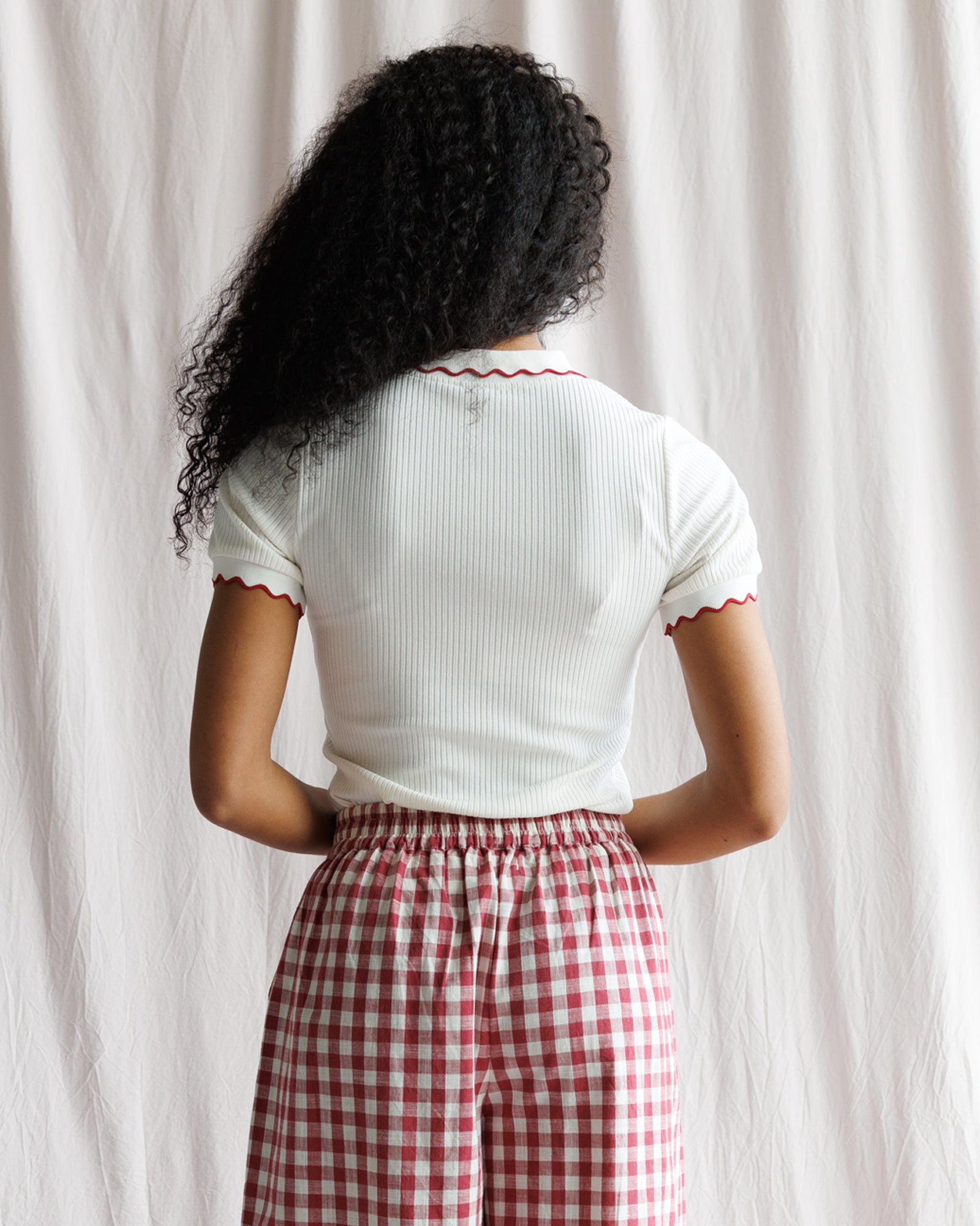 Person wearing a white ribbed top and red checkered pants against a light curtain background