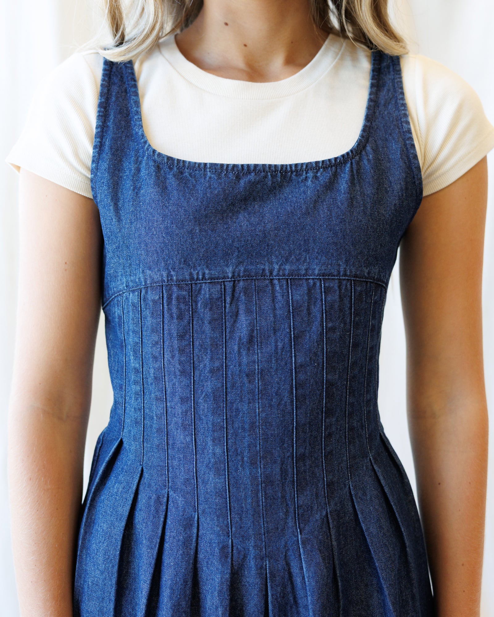 A person wearing a blue denim pinafore dress over a white short-sleeve t-shirt, shown from the neck down against a plain white background.