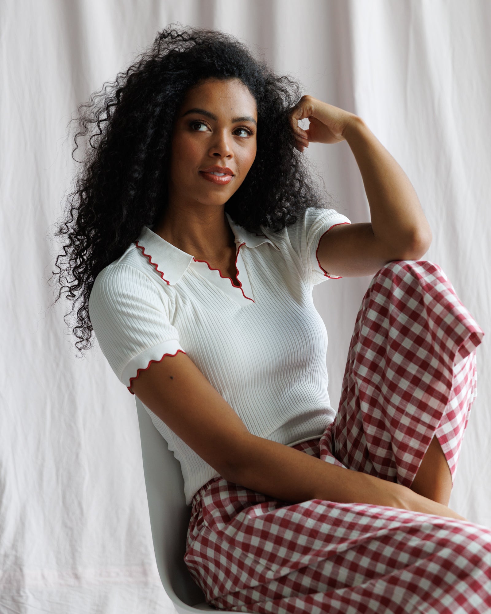Woman wearing a white top with red trim and red checkered pants sitting against a white curtain.