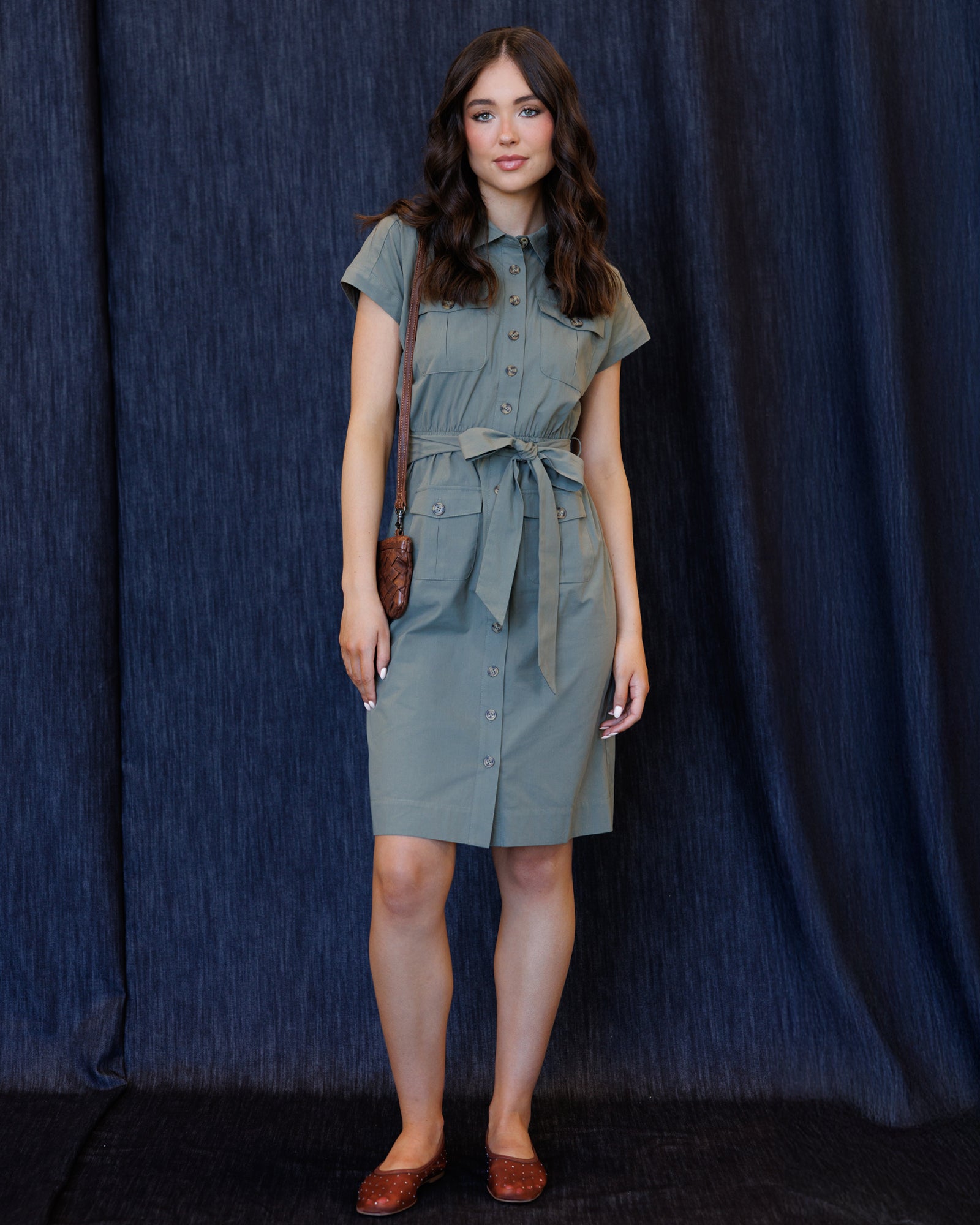 A woman modeling a sage green short-sleeve button-up shirt dress with a tie waist belt, standing against a dark blue textured curtain backdrop.
