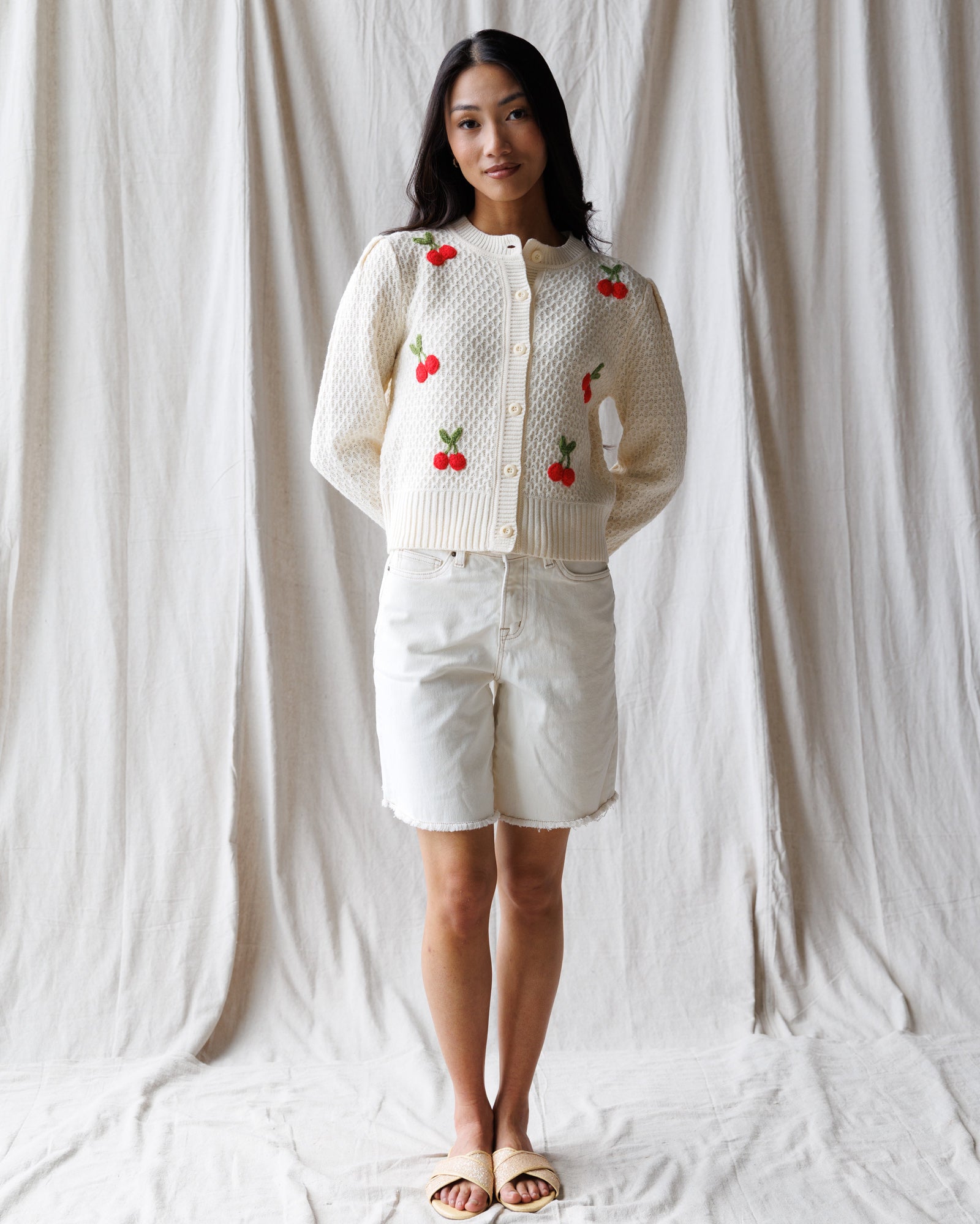 Woman wearing a white cardigan with red cherry patterns against a plain background