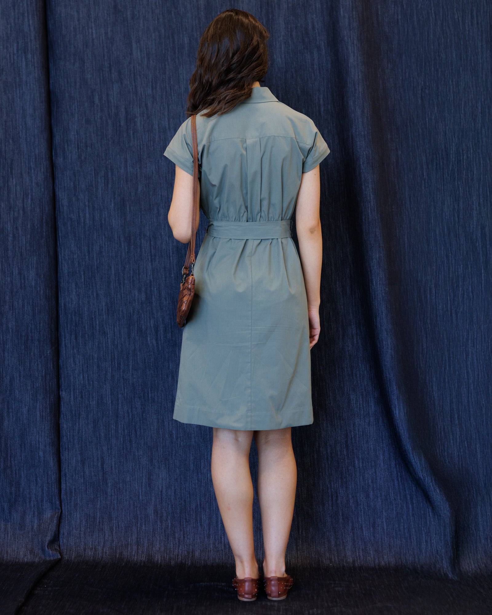 A woman stands with her back to the camera wearing a short-sleeve, collared, belted dress in a muted sage green. She is holding a small brown shoulder bag and wearing brown shoes, set against a dark blue textured fabric backdrop.