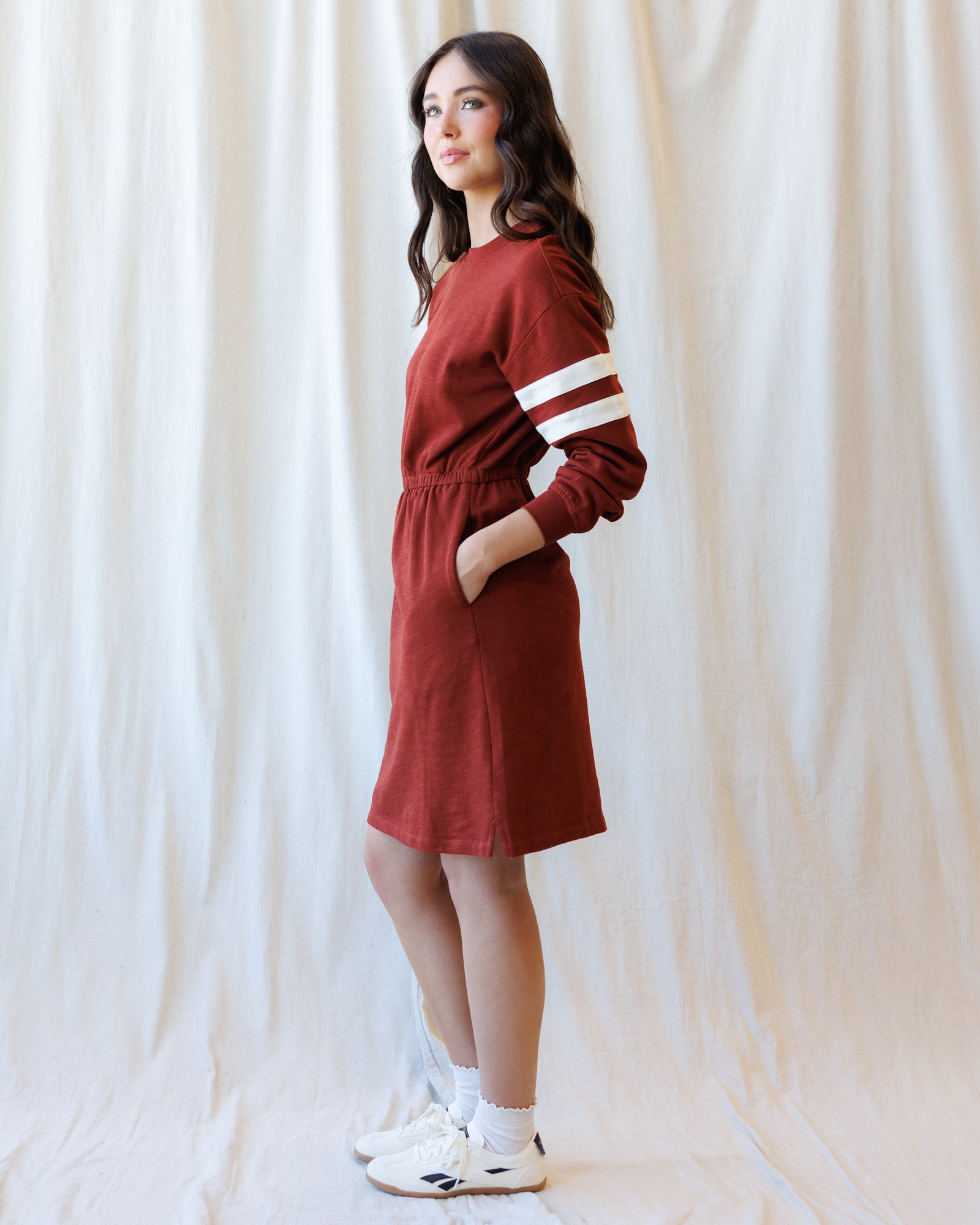 A woman modeling a rust-red long-sleeve dress with white stripes on the sleeves, standing against a draped white fabric backdrop.