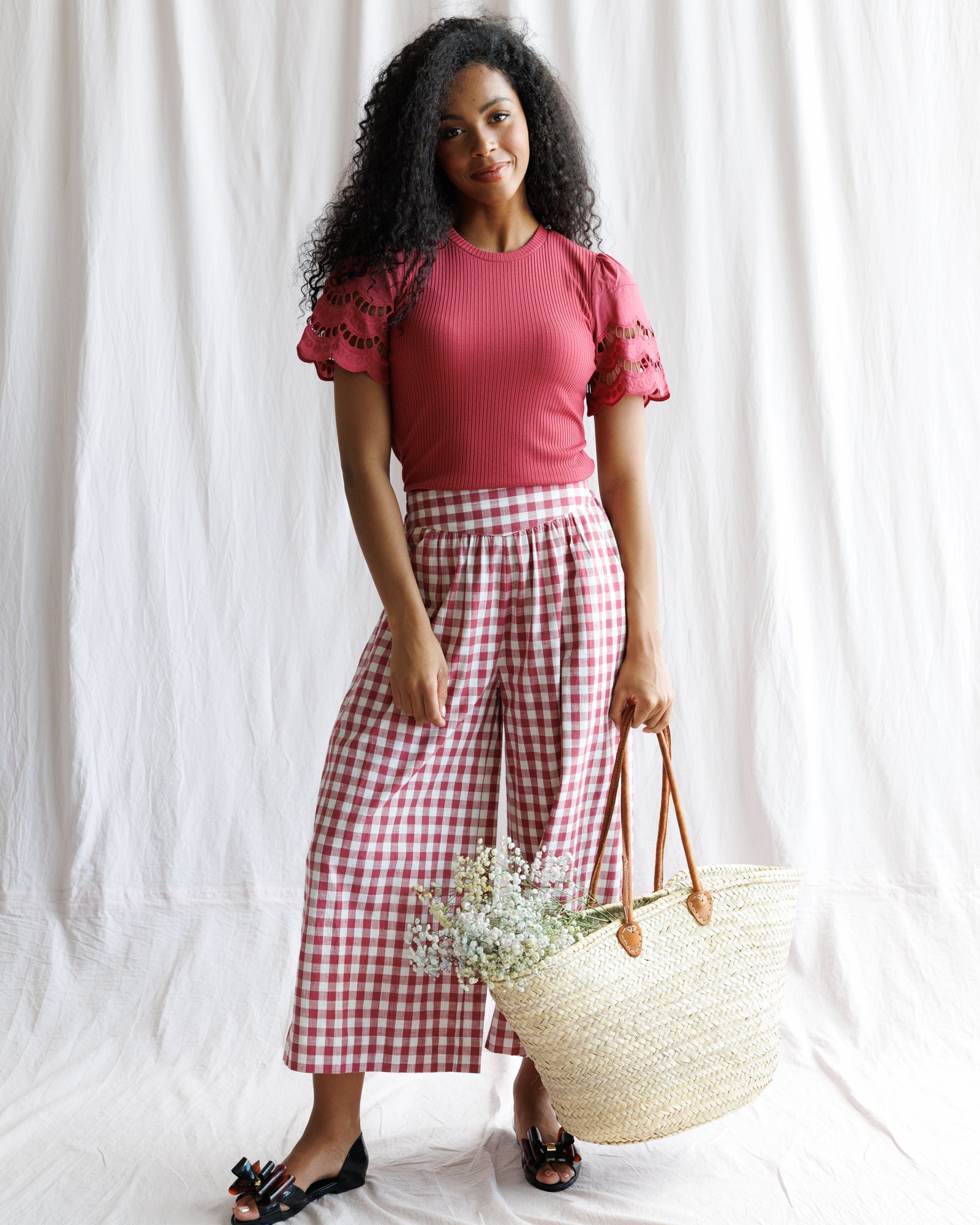Woman wearing a red top and pink checkered pants holding a straw bag against a white background