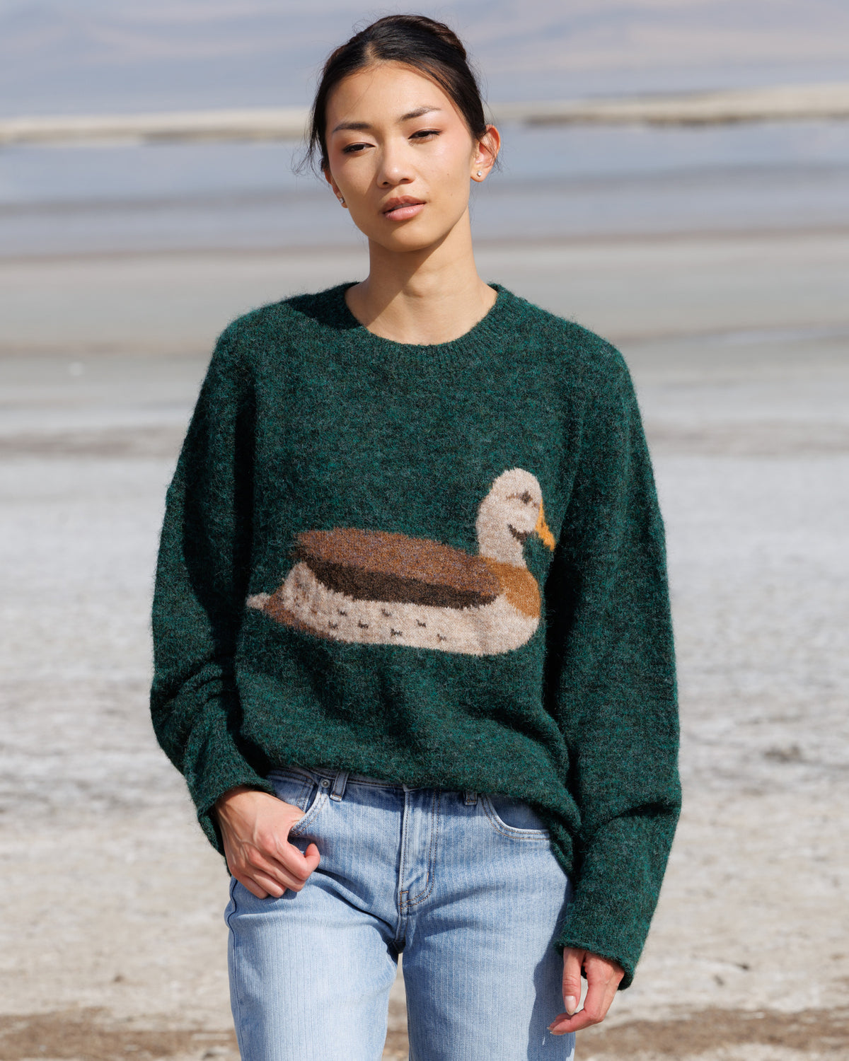 A woman wearing a dark green knit sweater with a large duck motif on the front, paired with light blue jeans, standing outdoors against a blurred natural background.