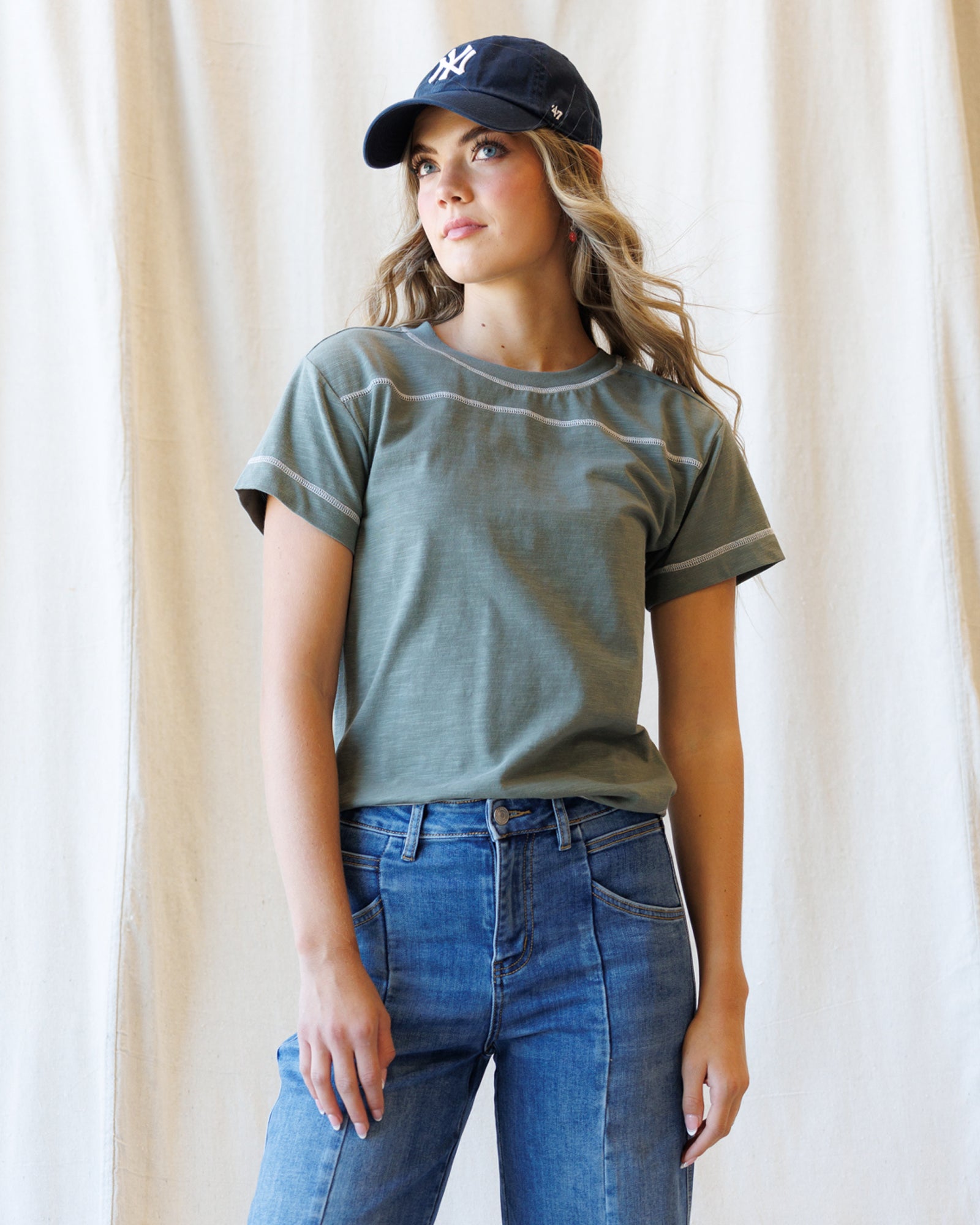 A woman wearing a short-sleeve crewneck t-shirt in a washed olive green color with subtle contrast stitching, paired with blue jeans and a navy baseball cap, standing against a neutral cream-colored curtain backdrop.