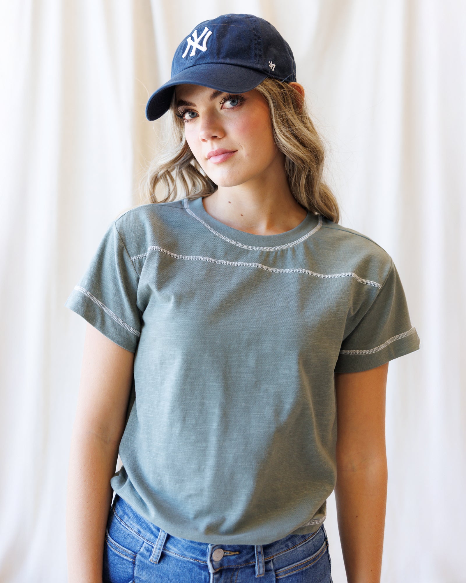 A woman wearing a faded sage green short-sleeve t-shirt with white contrast stitching, a navy blue New York Yankees baseball cap, and blue jeans, standing against a white curtain background.