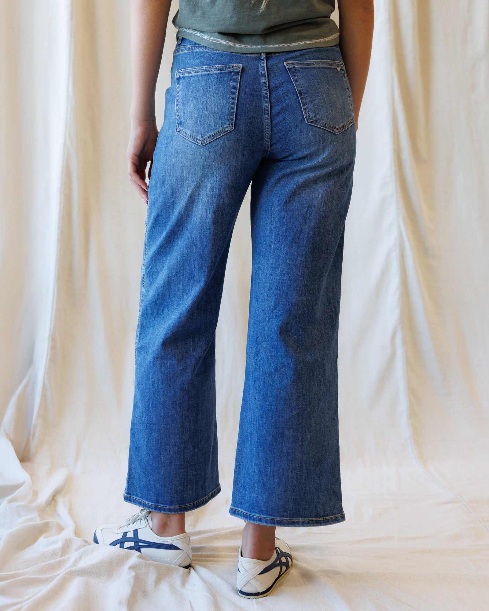 A person shown from the back wearing blue wide-leg cropped jeans, a gray top, and white sneakers with blue stripes, standing against a draped cream fabric backdrop.