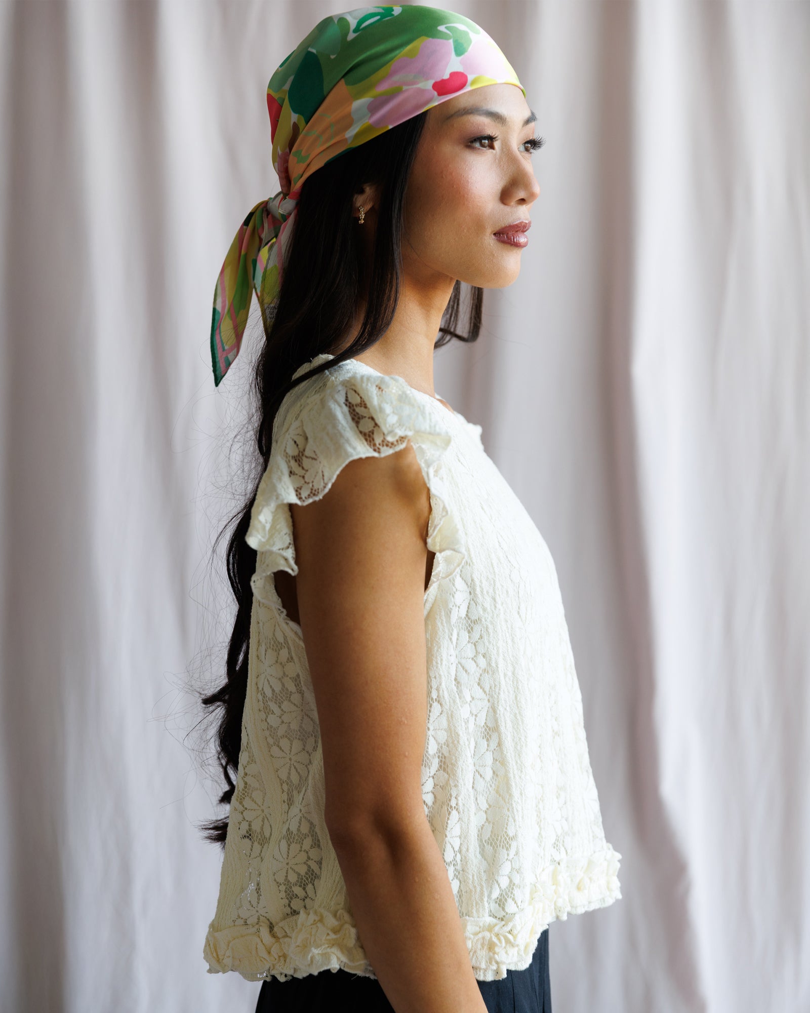 Woman wearing a colorful headscarf and white lace top against a plain background