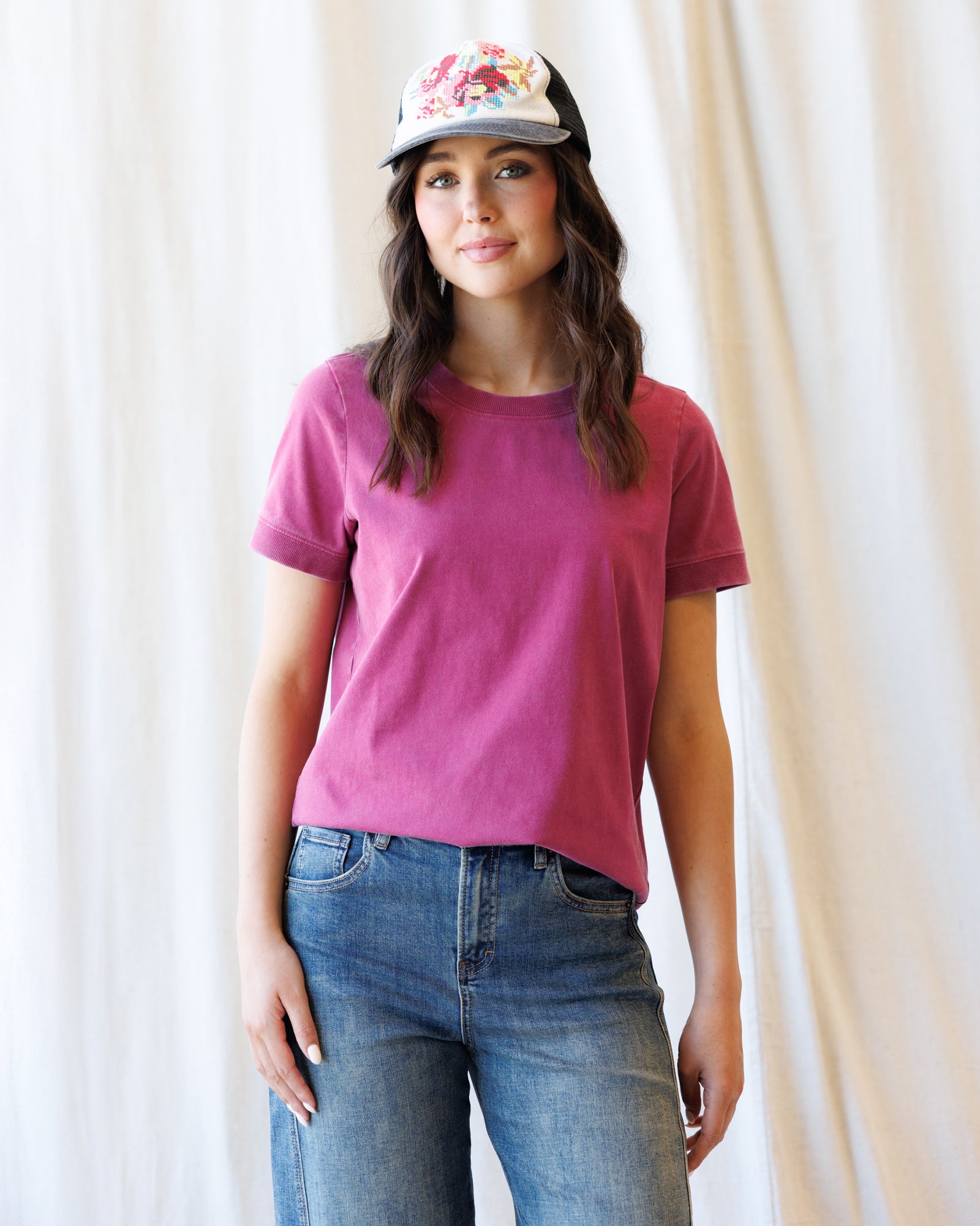 A woman wearing a short-sleeve crewneck t-shirt in a dusty magenta-pink color, paired with blue jeans and a baseball cap, standing against a draped cream fabric backdrop.