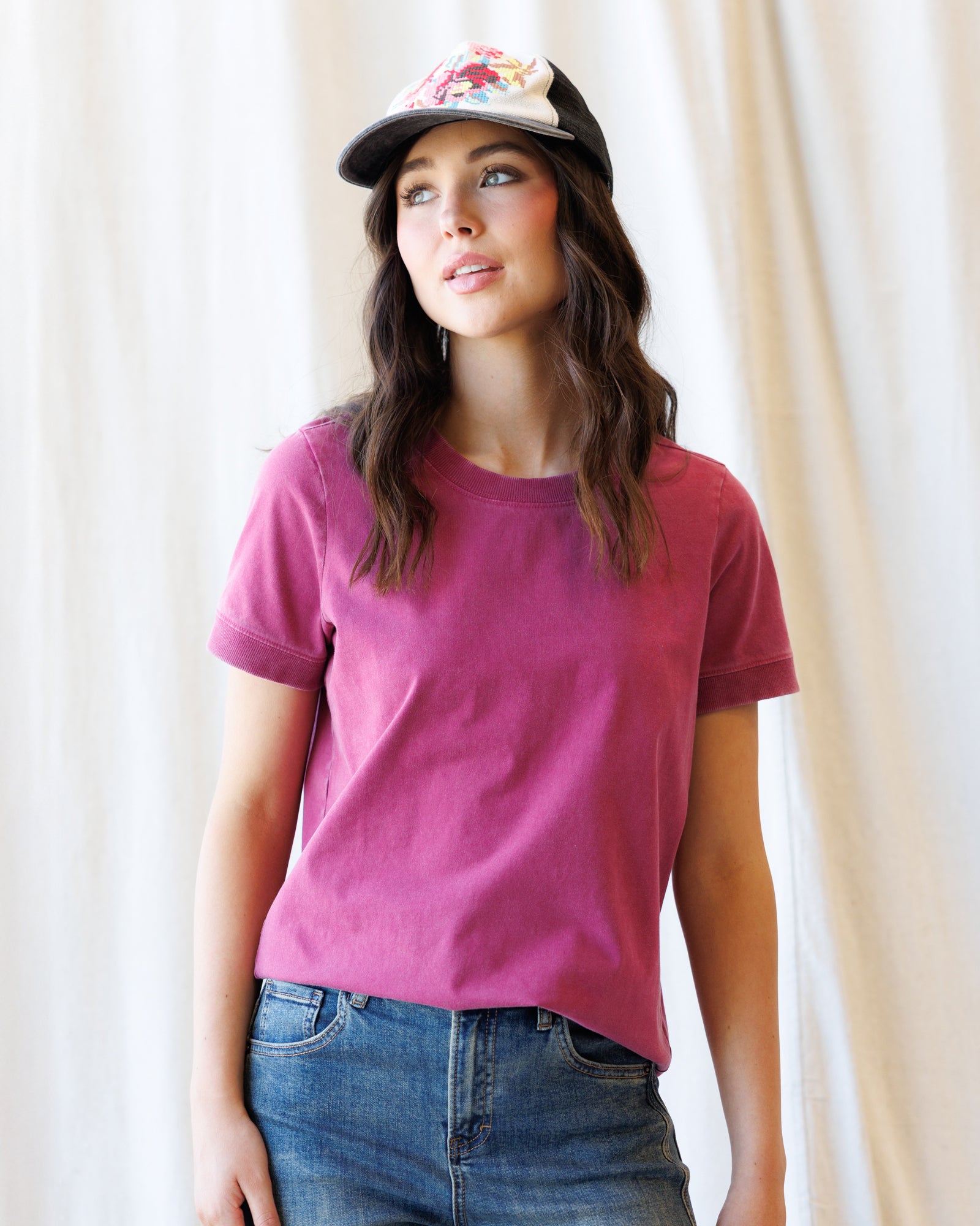 A woman wearing a short-sleeve crewneck t-shirt in a dusty magenta pink, paired with blue jeans and a baseball cap, standing against a soft beige curtain background.