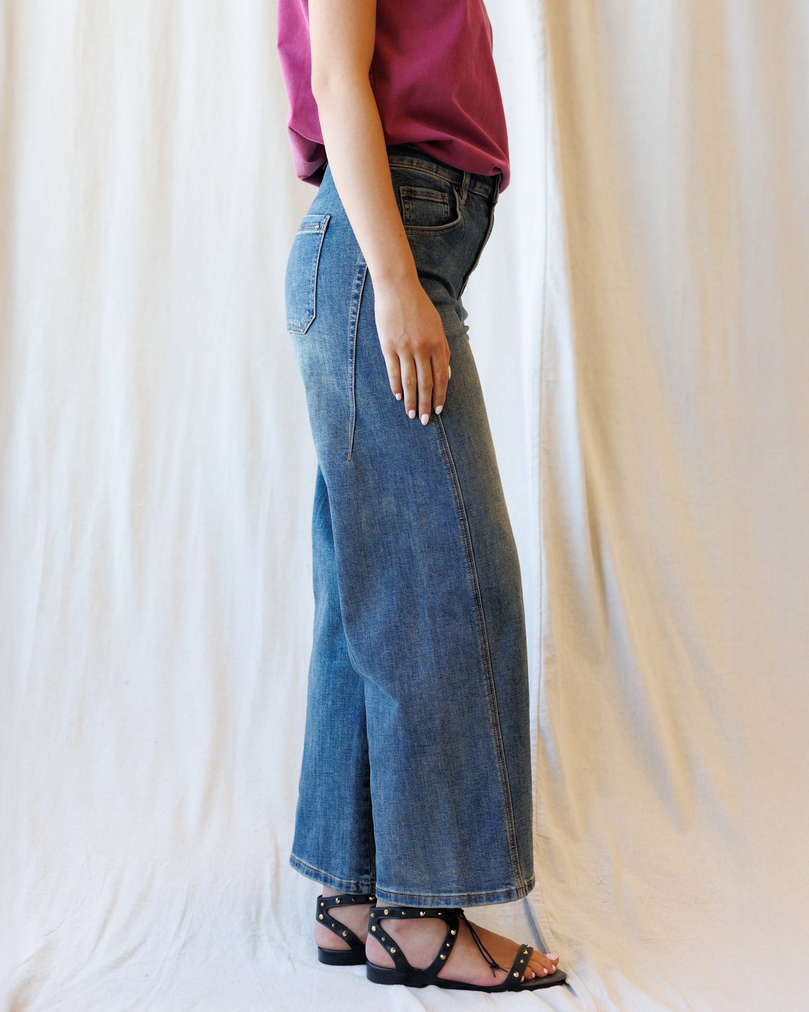 A person shown from the waist down wearing a pair of wide-leg blue denim jeans, paired with a magenta top and black studded sandals, standing against a draped white fabric backdrop.
